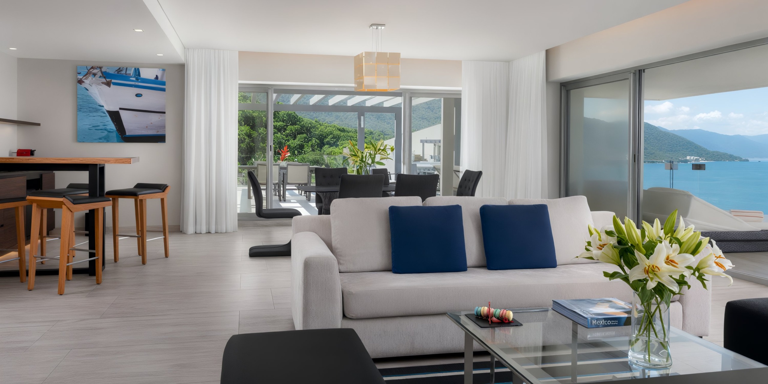 Experience a contemporary living space at the Hilton Vallarta Riviera All-Inclusive Resort, featuring a white sofa adorned with blue pillows and complemented by a sleek glass coffee table accented by fresh flowers. Large windows offer breathtaking views of the ocean and mountains, while the dining area is elegantly visible in the background.