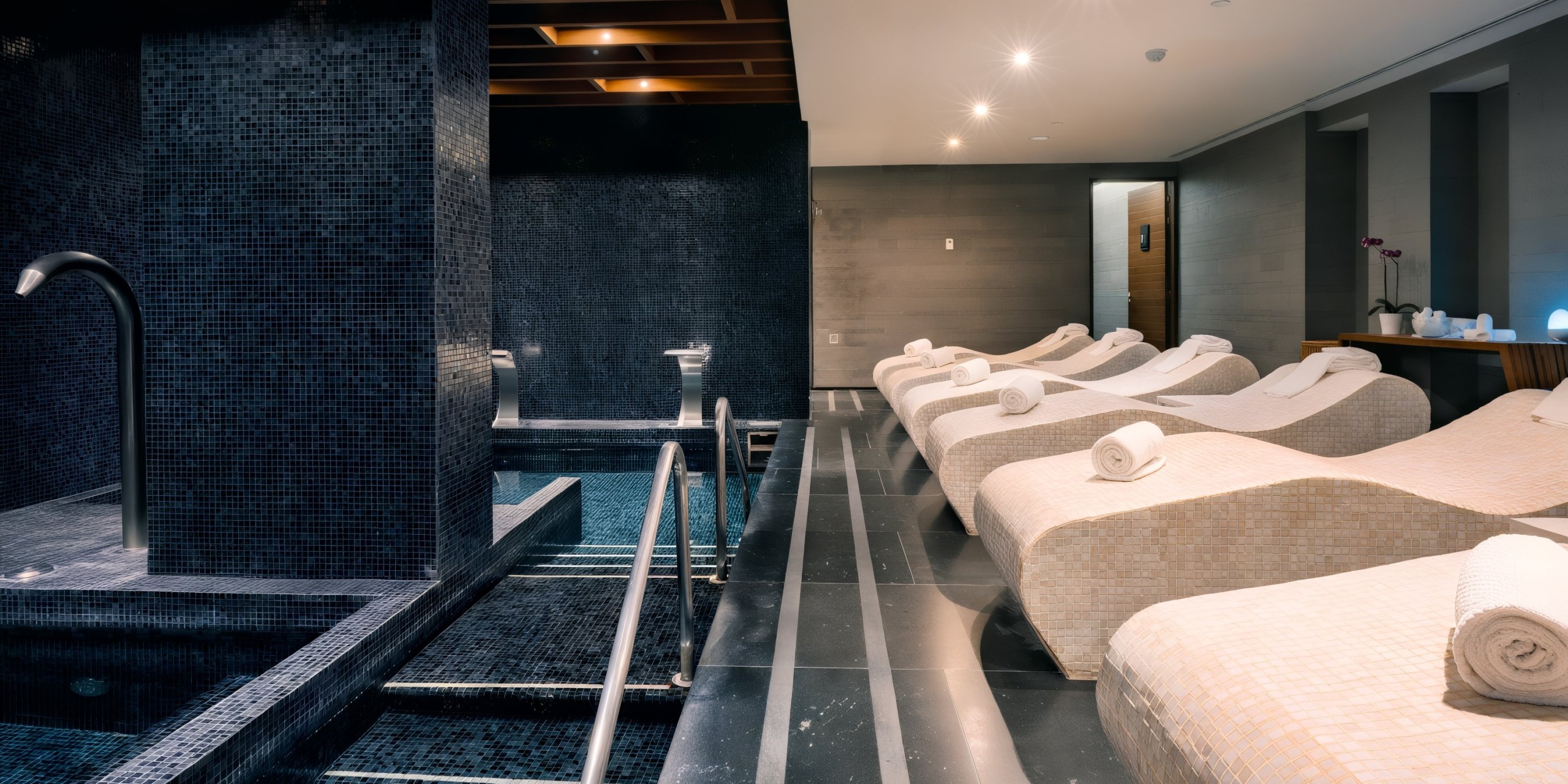 The Hilton Vallarta Riviera All-Inclusive Resort features a spa relaxation area with tiled lounge chairs, each adorned with a rolled towel. The serene atmosphere is enhanced by a mosaic-tiled pool and dim lighting.