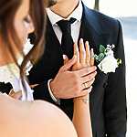 A bride and groom in formal attire hold hands, with their rings visible. The groom wears a black suit with a white rose boutonniere, and the bride wears a strapless gown, holding a bouquet.