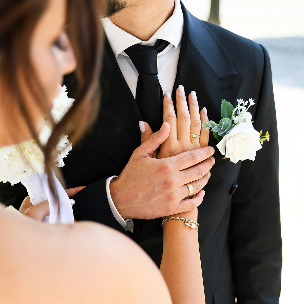 A bride and groom in formal attire hold hands, with their rings visible. The groom wears a black suit with a white rose boutonniere, and the bride wears a strapless gown, holding a bouquet.