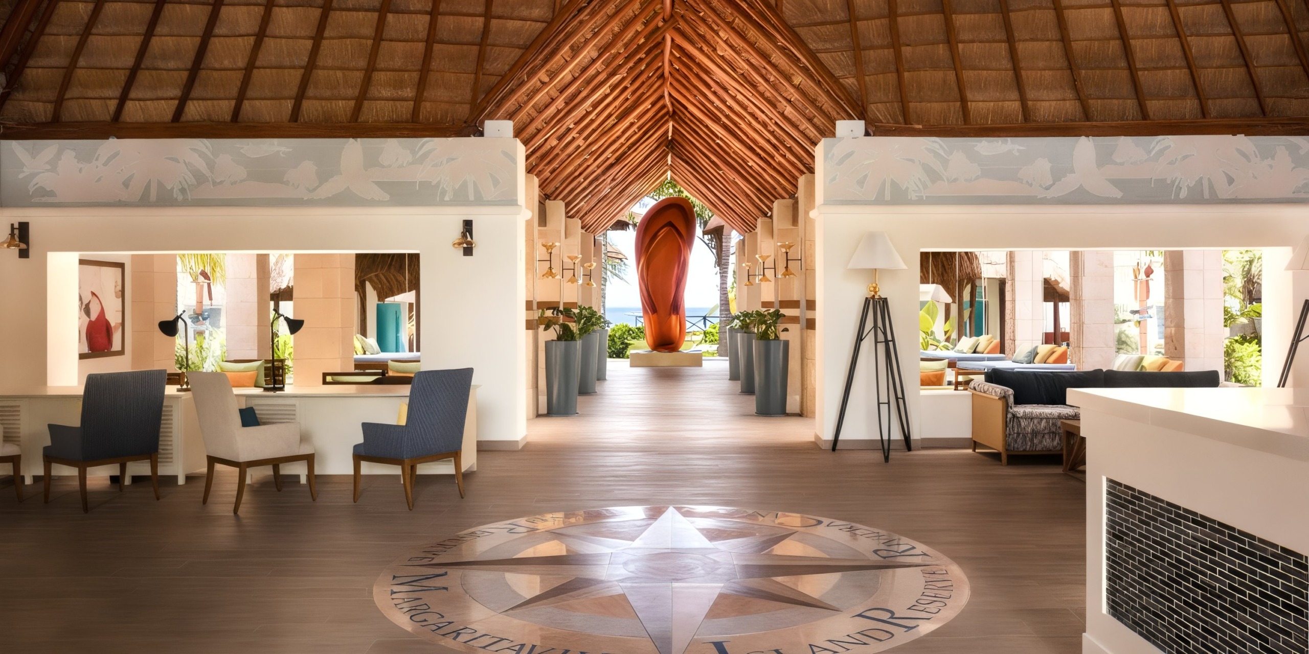 Lobby area at Margaritaville Island Reserve Resort Riviera Cancun with a high thatched ceiling, blue chairs, and a circular mosaic floor design. A large orange sculpture sits at the far end, offering glimpses of greenery and sky beyond.