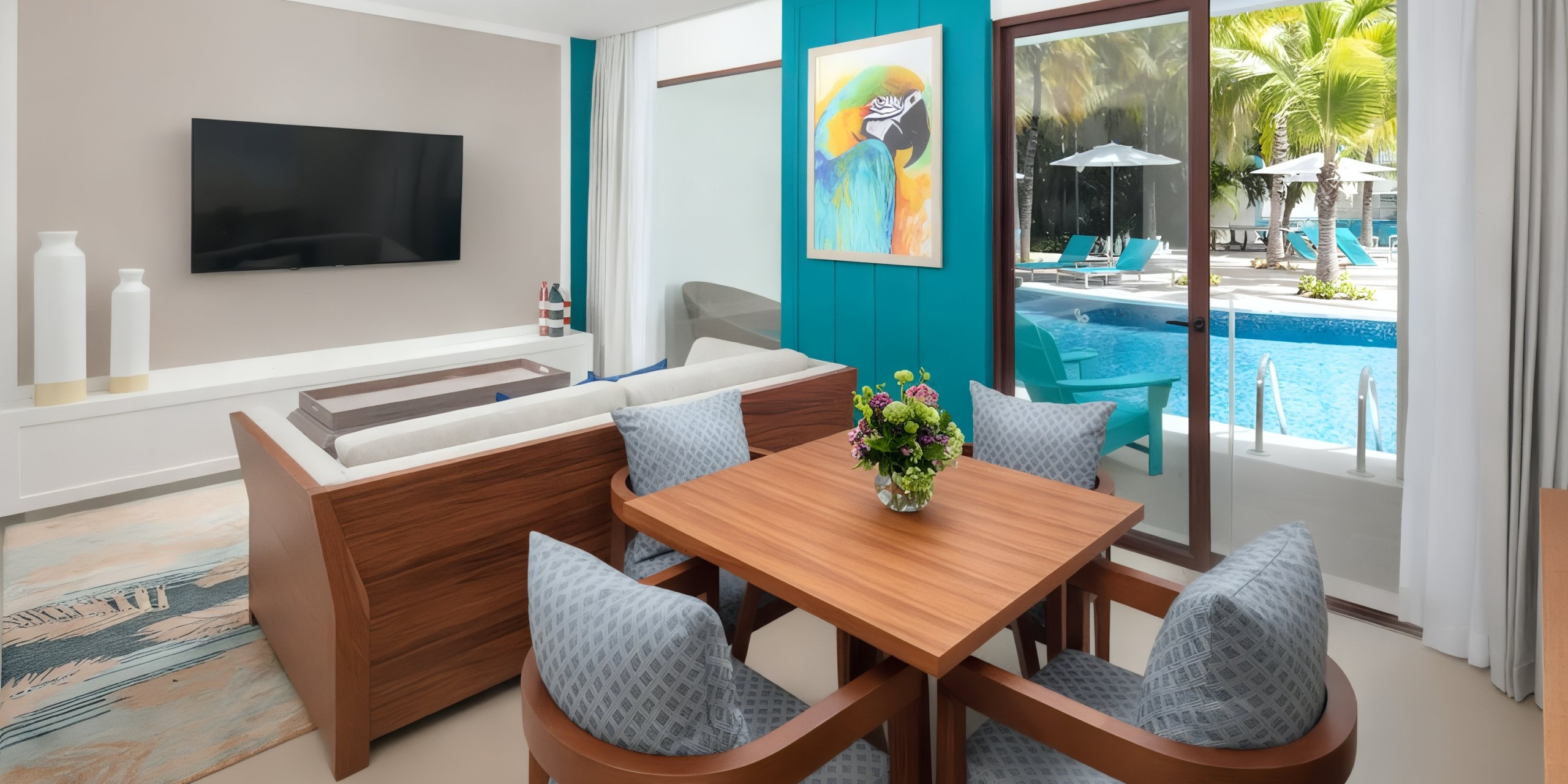 A contemporary room at Margaritaville Island Reserve Resort Riviera Cancun features a wooden dining table, cushioned chairs, a sofa, a wall-mounted TV, and vibrant parrot artwork. The patio door opens to reveal a pool view adorned with palm trees and loungers.