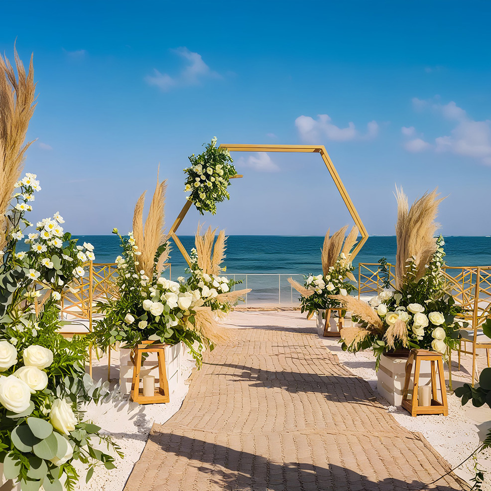 A beach wedding setup featuring a hexagonal wooden arch adorned with white flowers and greenery. Sandy path with floral arrangements on either side leads to the ocean.
