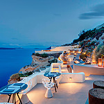 Outdoor seaside restaurant at dusk with tables set along a cliff, overlooking a calm ocean. The area is lit with ambient lights, creating a cozy atmosphere.