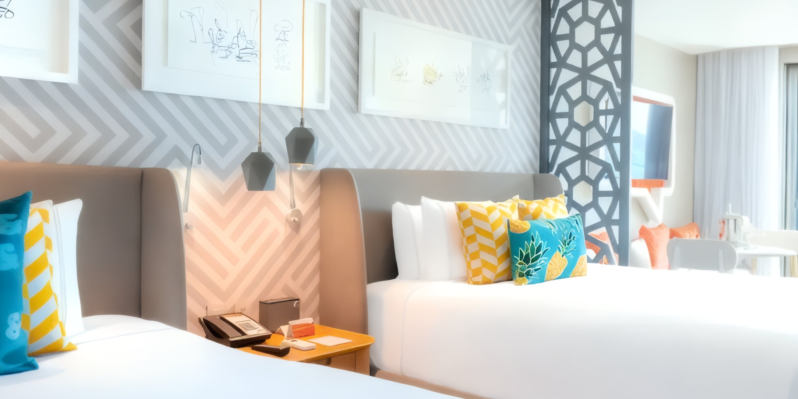 Experience the vibrant ambiance of Nickelodeon Hotels & Resorts Riviera Maya, featuring a well-lit hotel room with two cozy beds, adorned with decorative pillows. Geometric wall art adds a touch of elegance, while hanging pendant lights create a warm and inviting atmosphere.