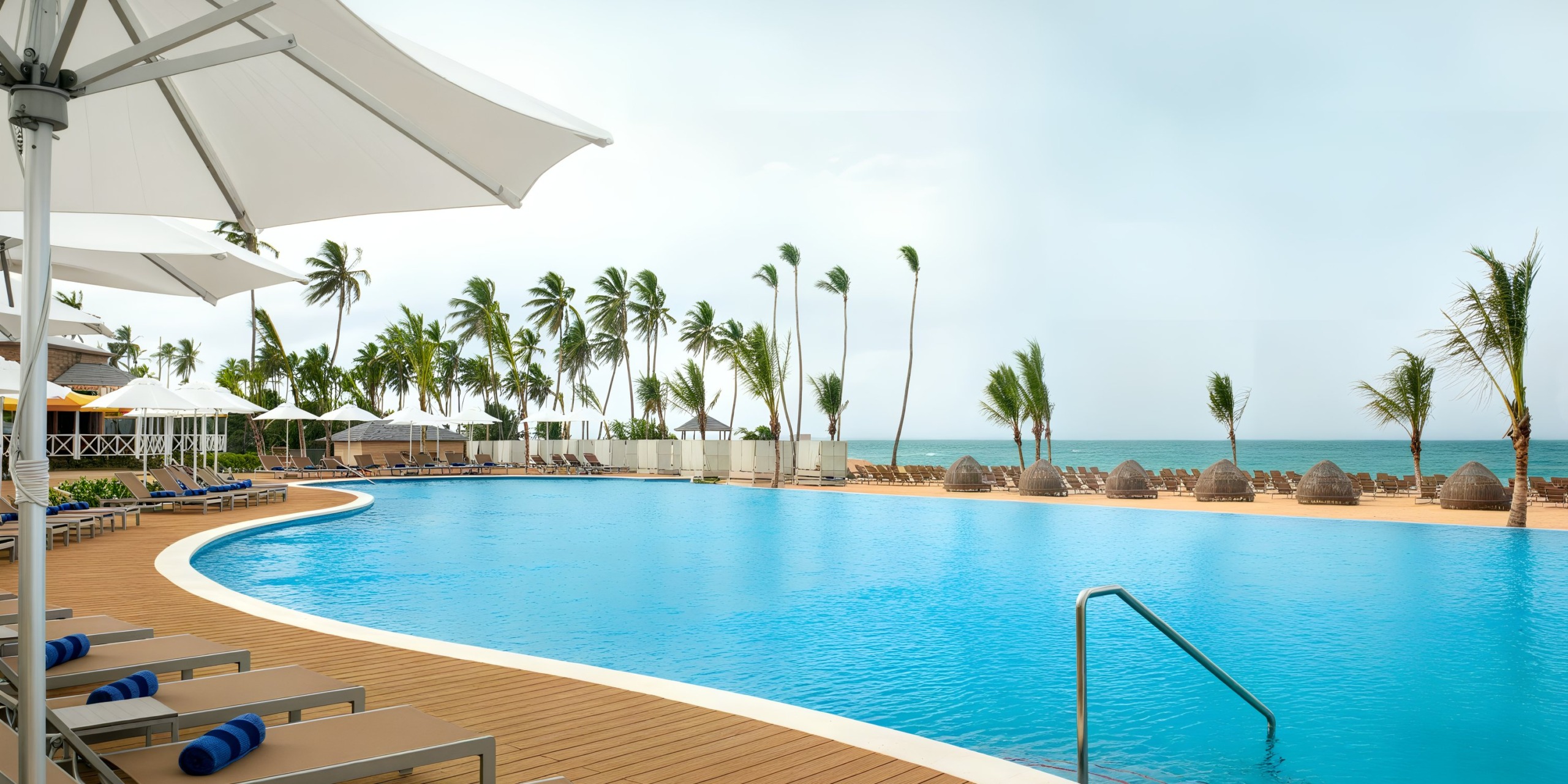At Nickelodeon Hotels & Resorts Riviera Maya, guests can enjoy a spacious, curved swimming pool complete with lounge chairs and umbrellas, all set against the backdrop of a beach lined with palm trees under a clear sky.