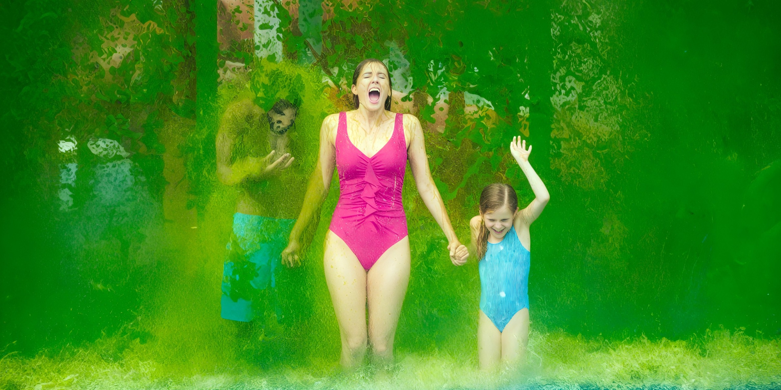 A woman and a child in swimsuits stroll hand in hand through a refreshing green water spray at Nickelodeon Hotels & Resorts Riviera Maya, both beaming with joy, while a person is partially visible in the background.