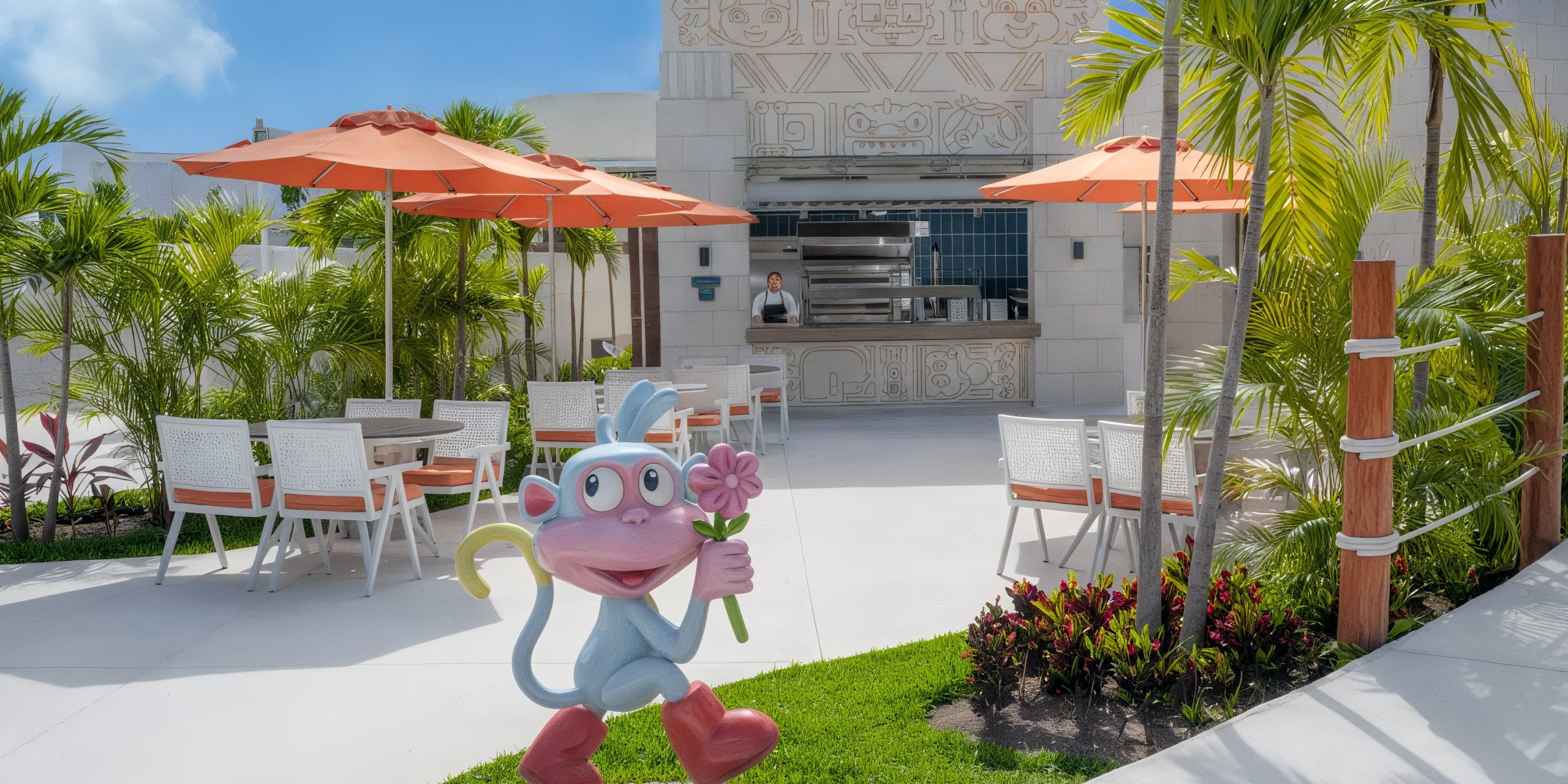 The outdoor seating area at Nickelodeon Hotels & Resorts Riviera Maya features tables, chairs, and umbrellas. A cartoon monkey statue holding flowers stands in the foreground, surrounded by tropical plants that adorn the space.
