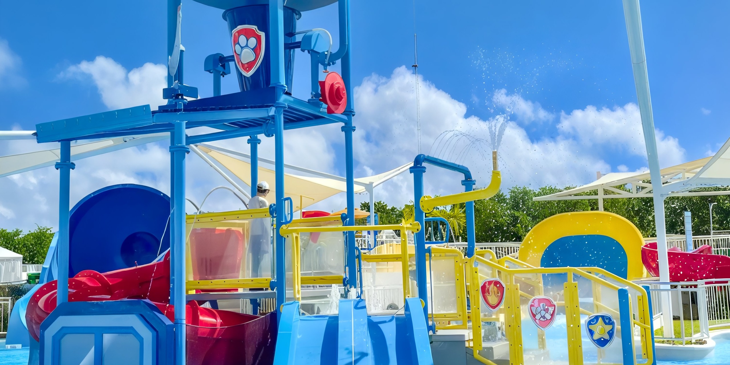The Nickelodeon Hotels & Resorts Riviera Maya offers a children's water play area with colorful slides, climbing structures, and water sprays set against a blue sky.