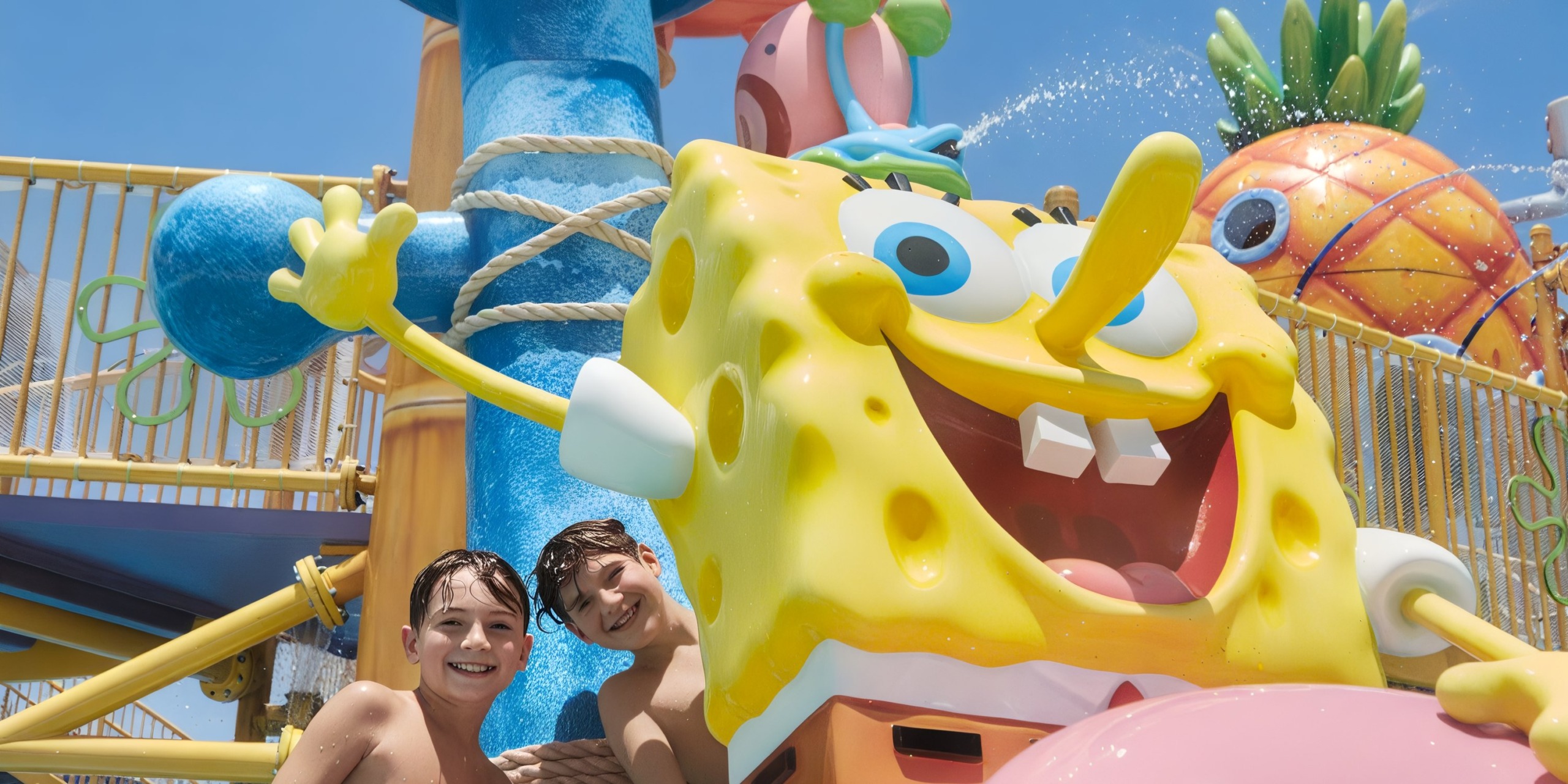 Two kids smiling next to a large SpongeBob SquarePants statue at Nickelodeon Hotels & Resorts Riviera Maya, with colorful structures and splashes of water in the background.