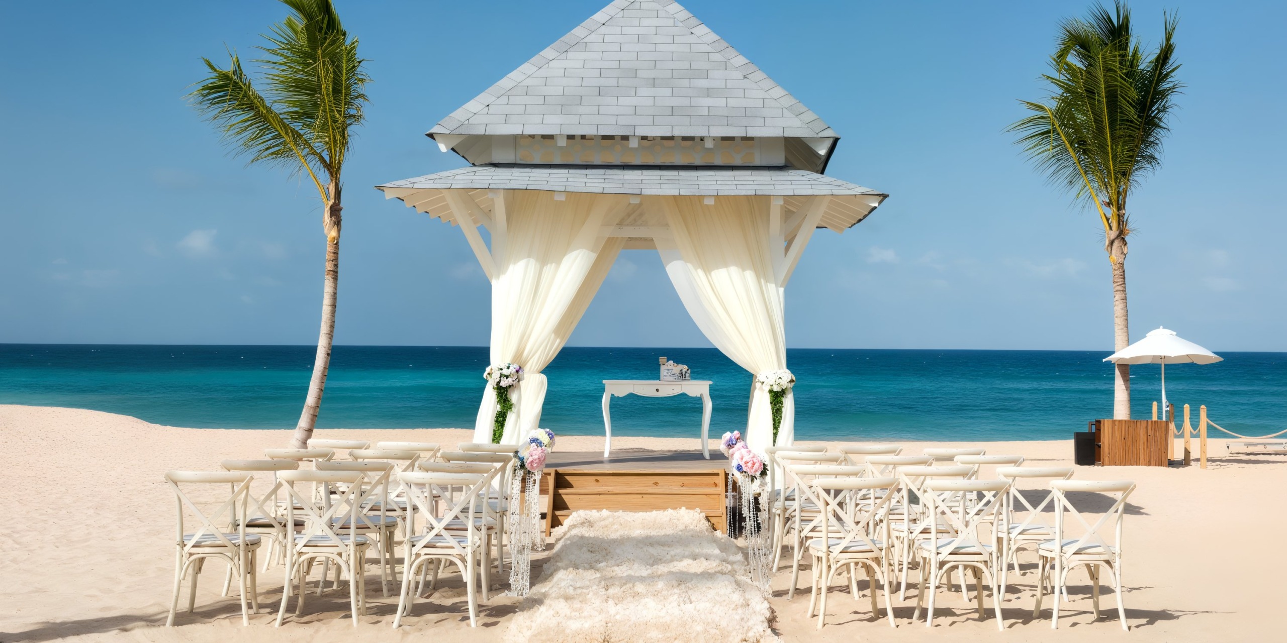 Experience a beachside wedding setup at Nickelodeon Hotels & Resorts Riviera Maya, featuring white chairs arranged to face a draped wooden gazebo on sandy shores, with the clear blue ocean as a stunning backdrop and palm trees framing the idyllic scene.
