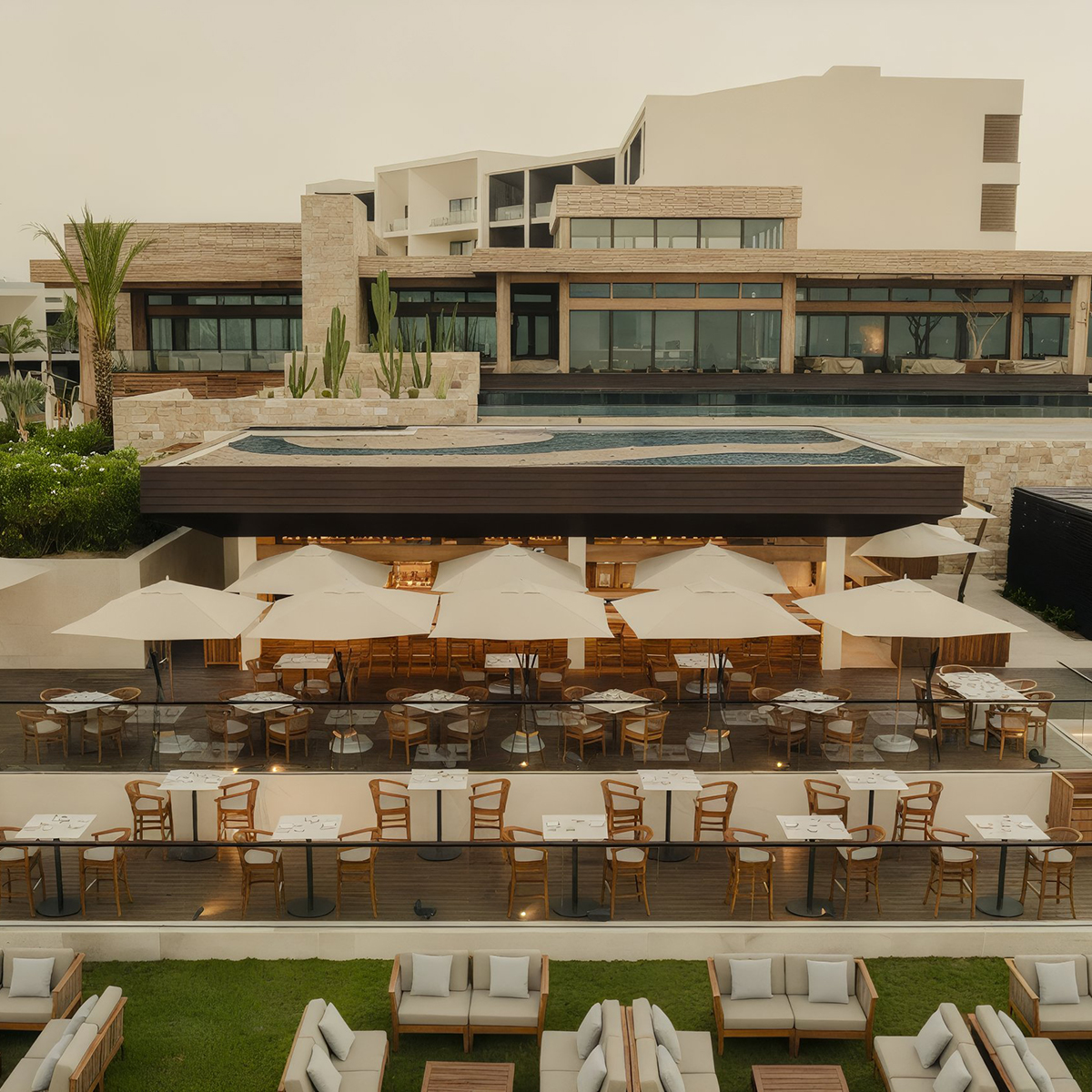 Outdoor restaurant with wooden tables and chairs under large umbrellas. Above is a rooftop pool. The building has modern architecture with stone and glass features.