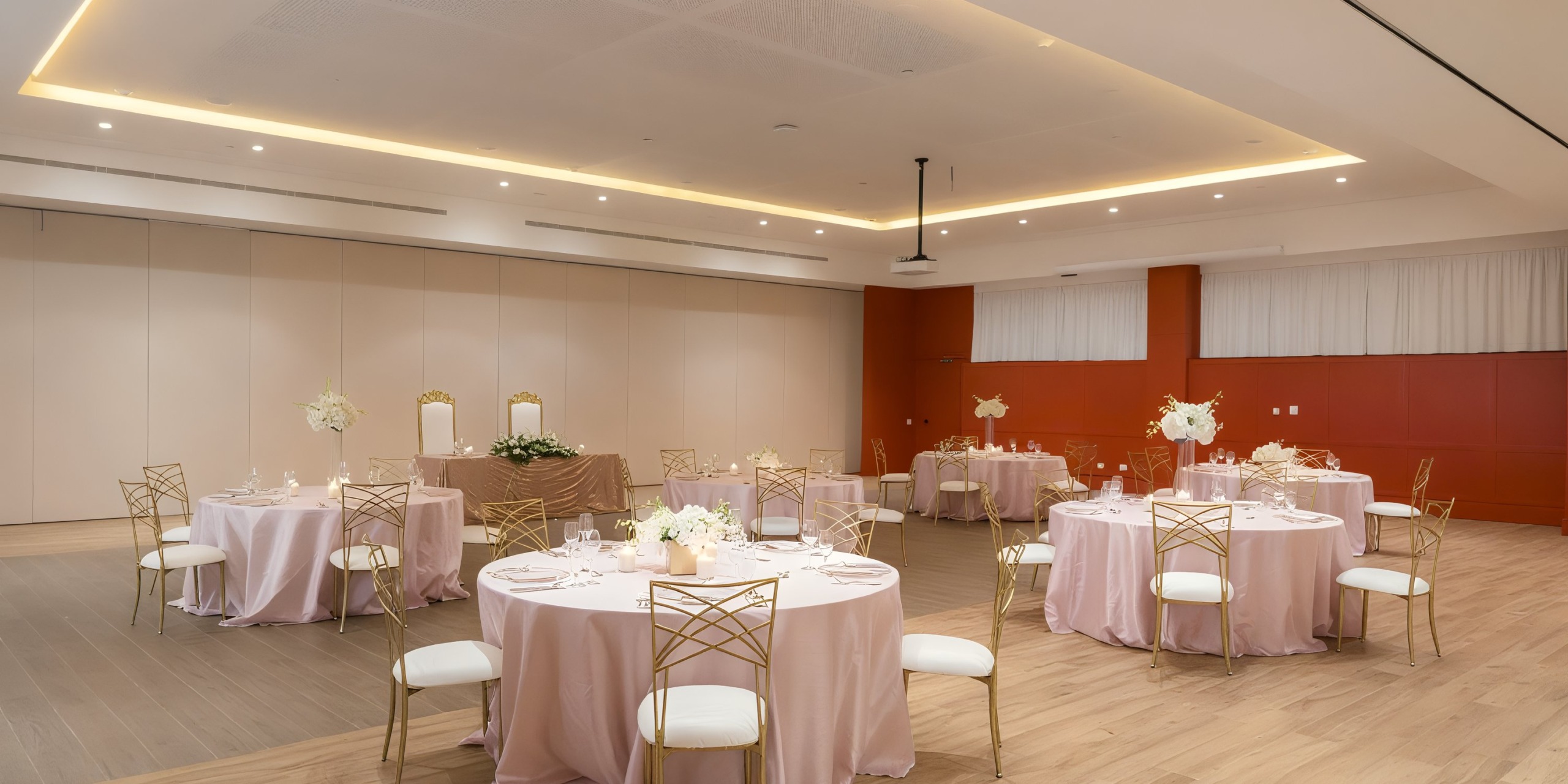 The Ocean Eden Bay banquet hall features round tables adorned with pink tablecloths, elegant chairs, and charming floral centerpieces. The room is enhanced with wooden flooring and soft, warm lighting for a spacious and inviting atmosphere.