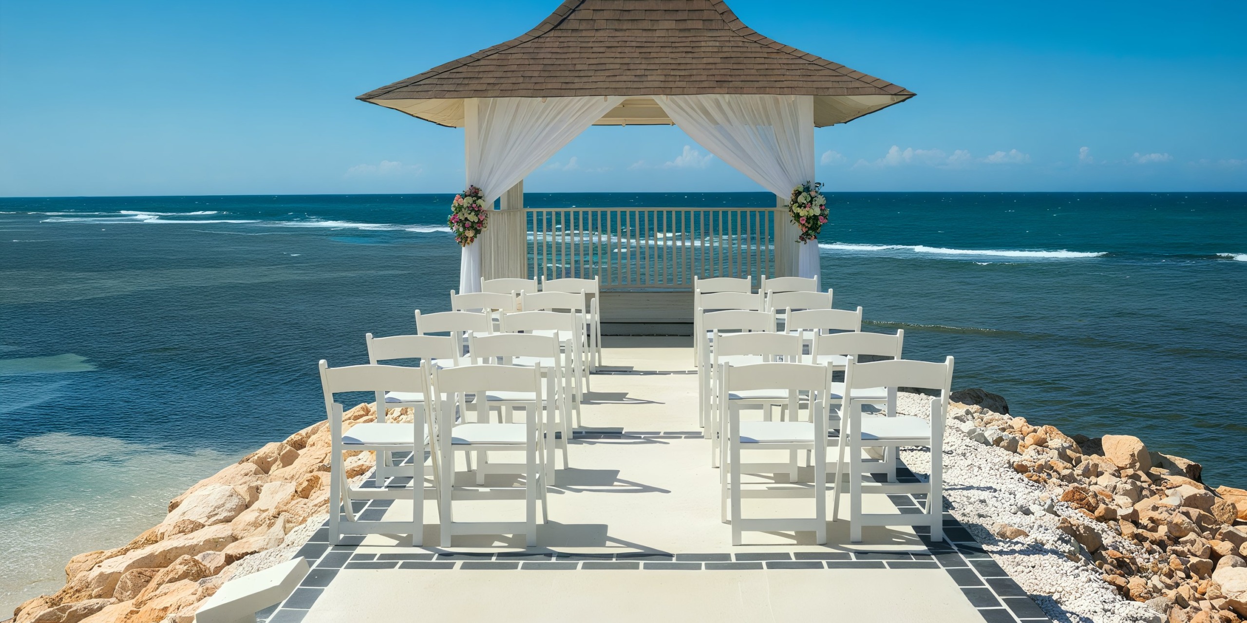A seaside wedding setup at Ocean Eden Bay features white chairs facing a gazebo beautifully adorned with flowers, all overlooking the ocean beneath a clear blue sky.
