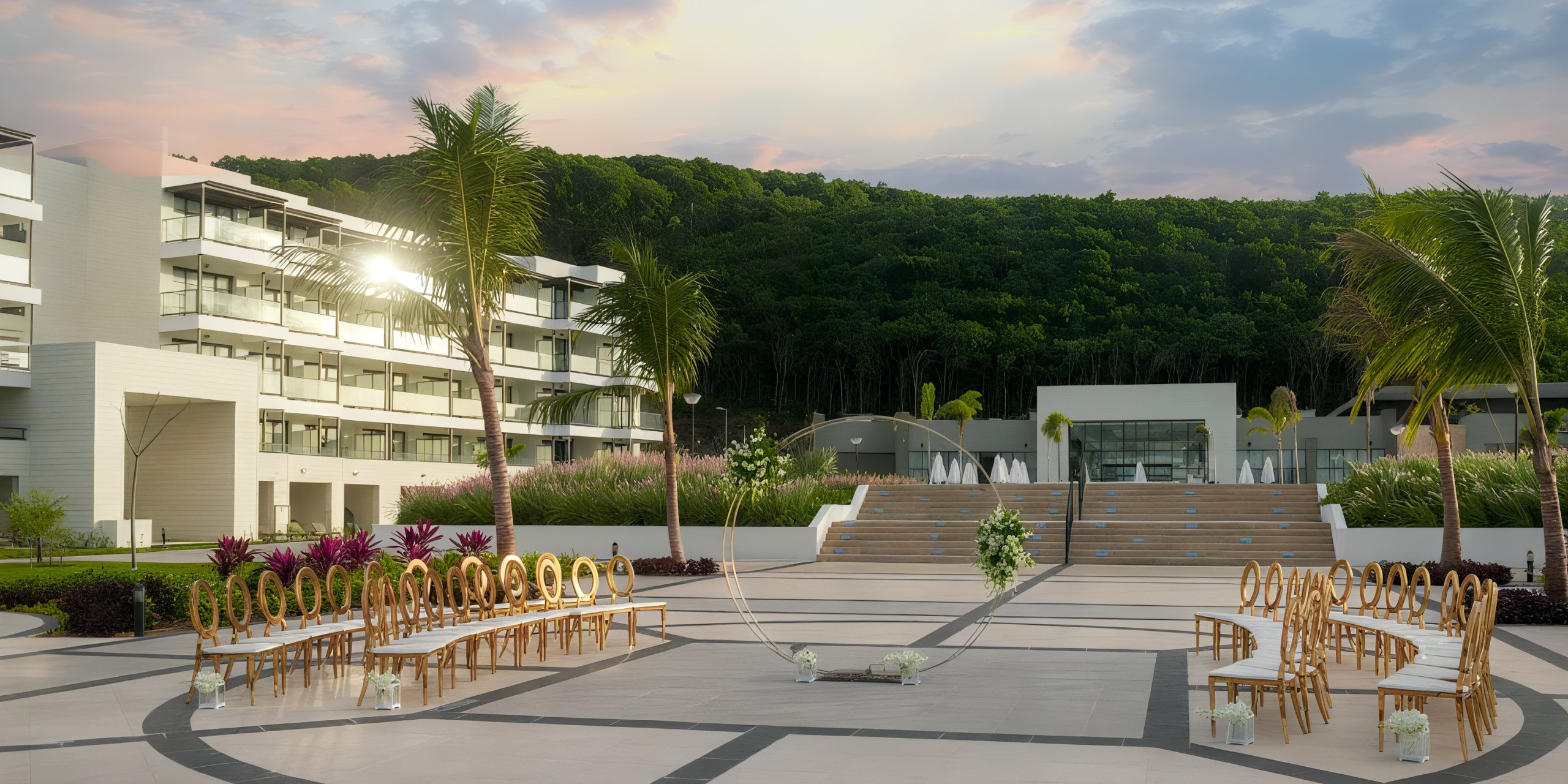 Ocean Eden Bay offers an outdoor wedding venue featuring contemporary architecture, arranged seating, and a floral arch. Steps guide you to a wooded area beneath a pink and blue sky.