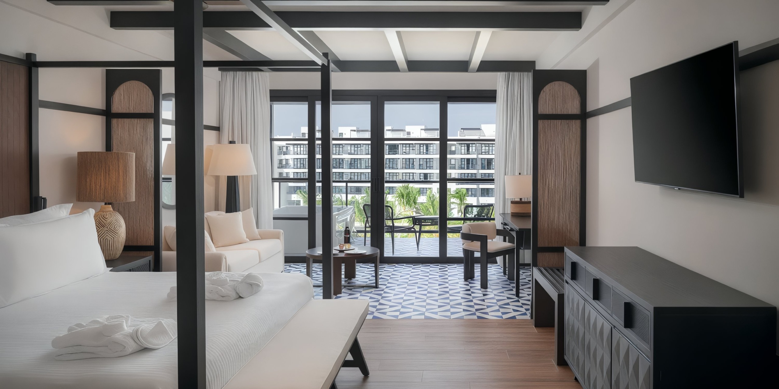 Stay in a contemporary room at Ocean Eden Bay, featuring a four-poster bed, comfortable sofa, and a large TV. Enjoy the balcony view of palm trees and nearby buildings.