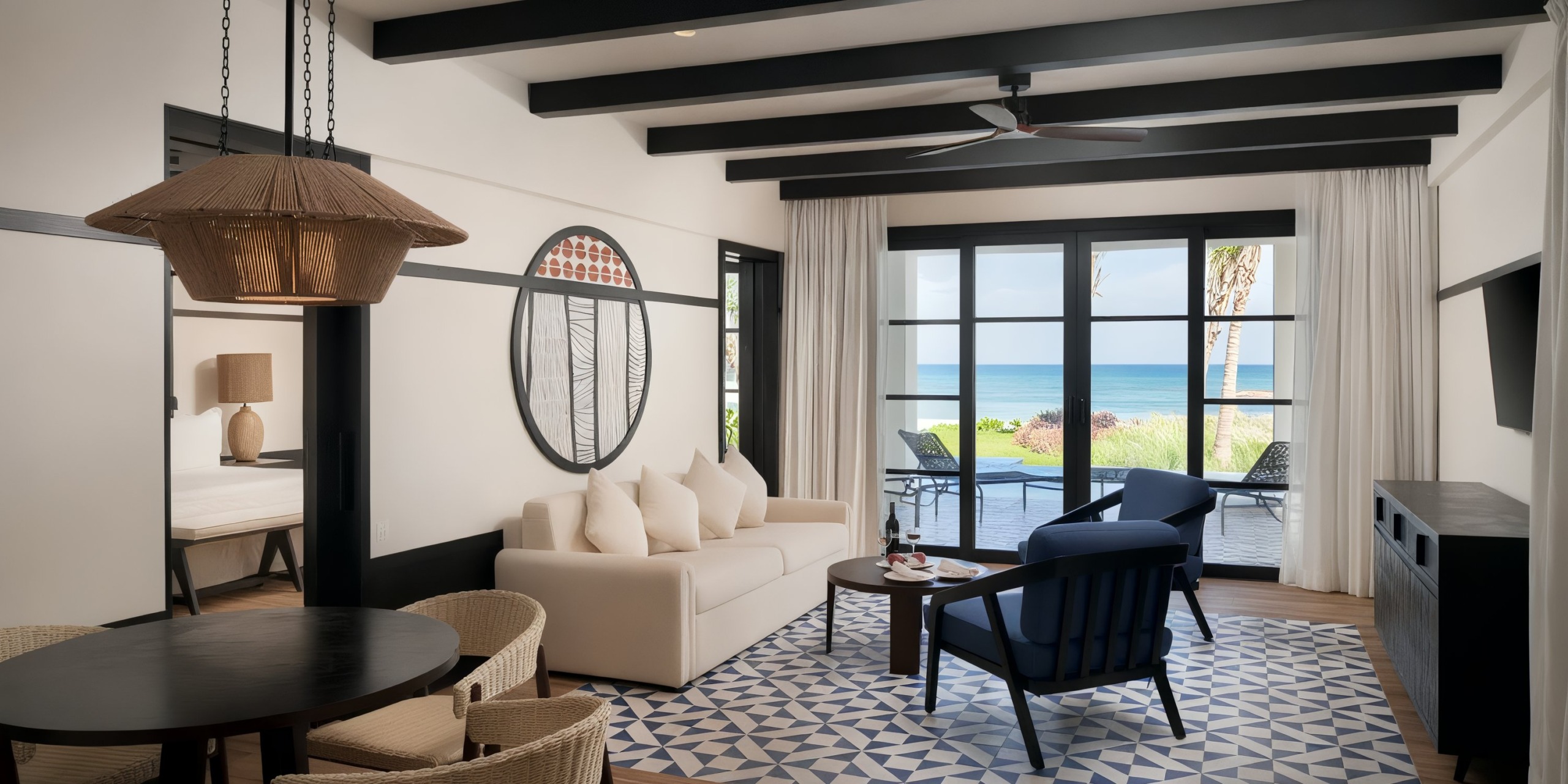 Explore the Ocean Eden Bay living room, showcasing a beige sofa paired with blue armchairs and a geometric rug. Large windows offer breathtaking views of the ocean, while decor features include a round wall art piece and a woven pendant light to complete the coastal ambiance.