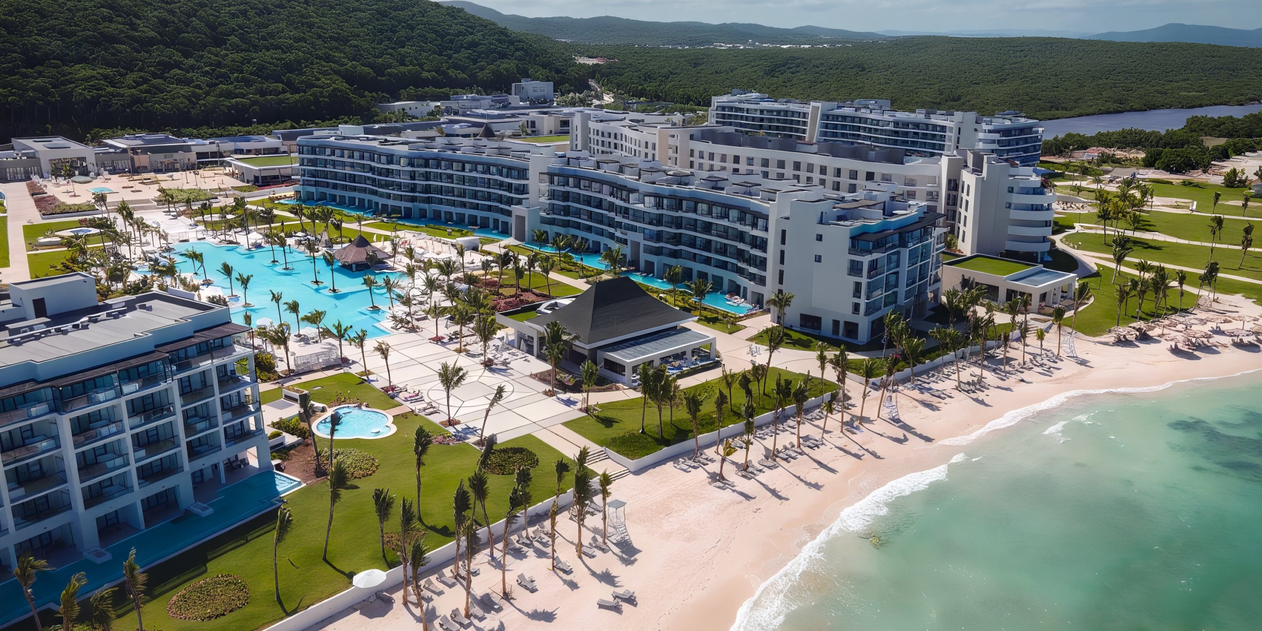 An aerial view of Ocean Eden Bay, a spacious beachfront resort featuring multiple buildings, a swimming pool, and palm trees lining the sandy beach, with hills and lush greenery visible in the background.