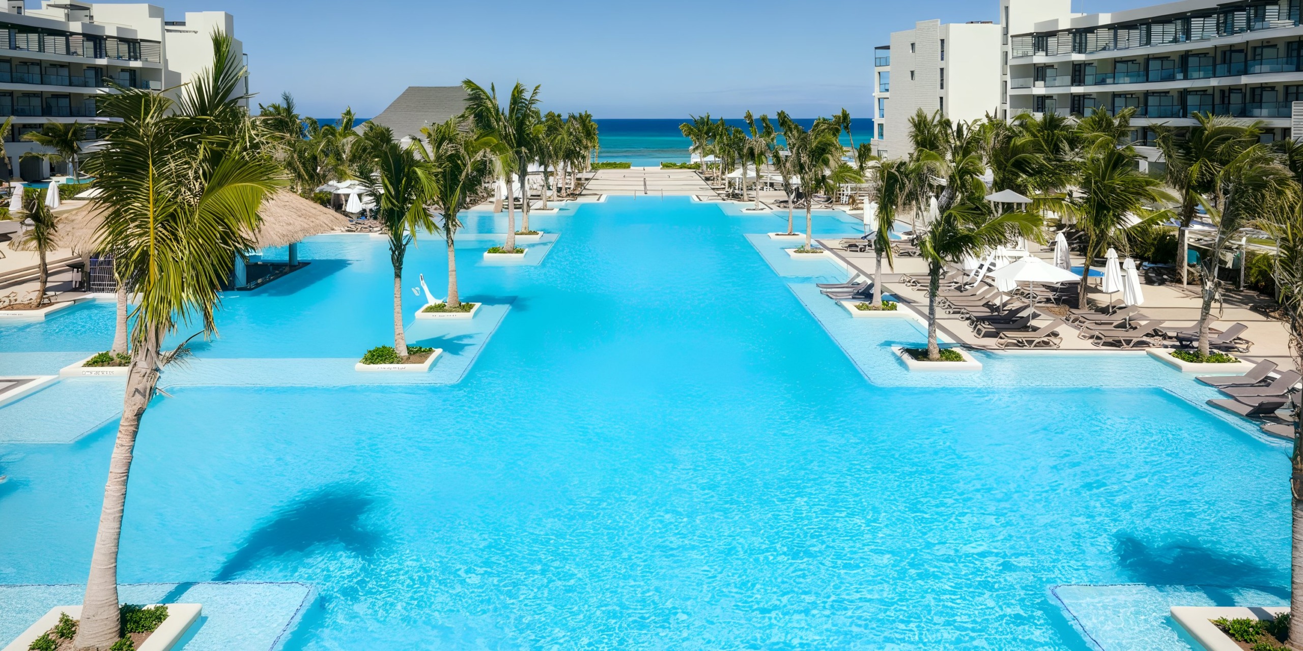 The Ocean Eden Bay features a spacious resort pool flanked by palm trees and lounge chairs, encircled by contemporary buildings, with an ocean view in the background.