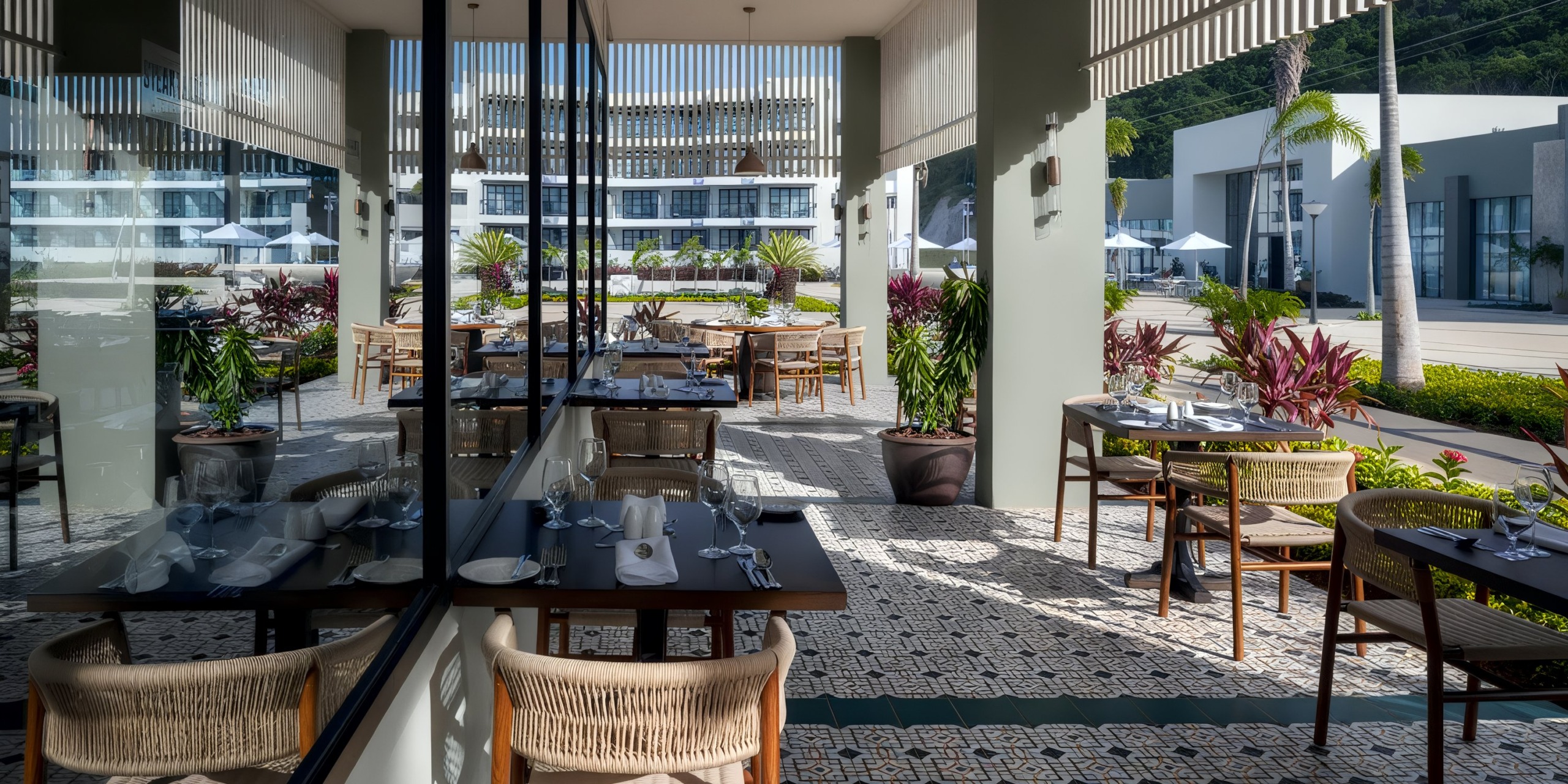 The outdoor restaurant seating area at Ocean Eden Bay boasts modern decor, complete with wooden tables and chairs, patterned floor tiles, and lush tropical plants. Large glass panels beautifully reflect the setting.