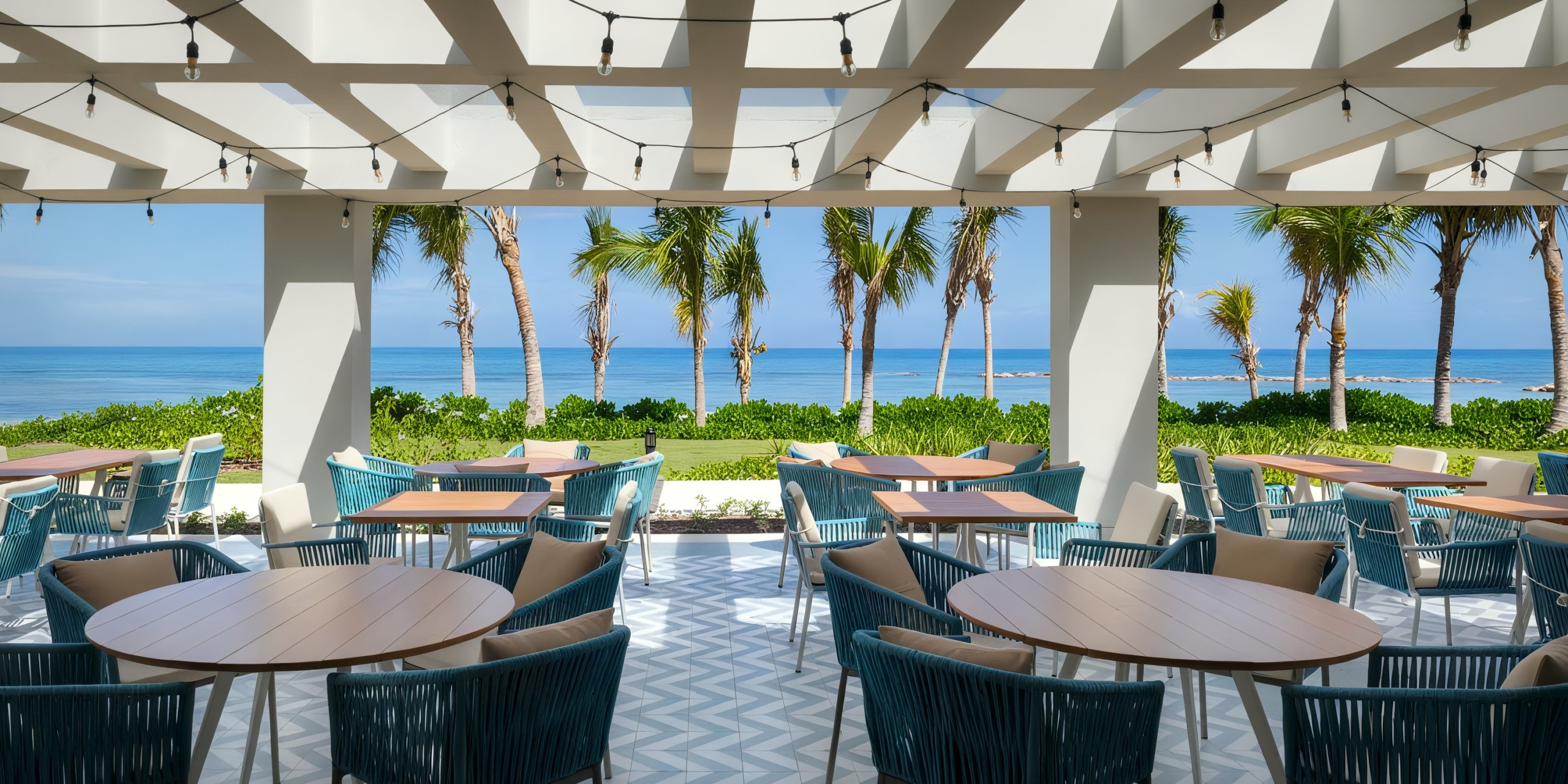The outdoor dining area at Ocean Eden Bay features round tables and blue chairs under a pergola, offering views of palm trees and a beach.