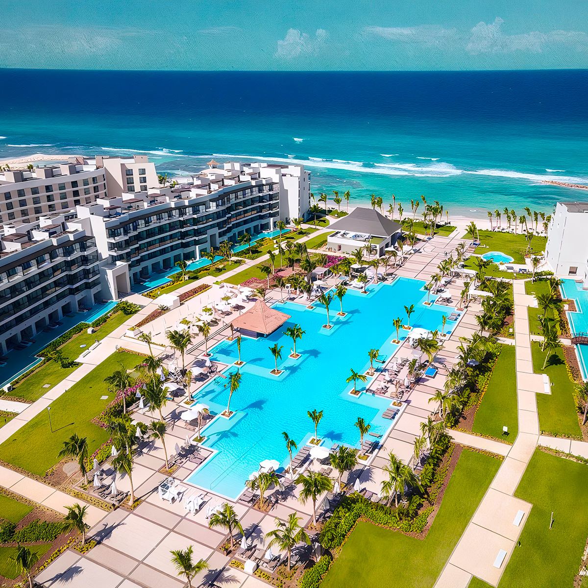 Aerial view of Ocean Eden Bay, a seaside resort featuring a large pool, palm trees, and surrounding modern buildings with the ocean in the background.
