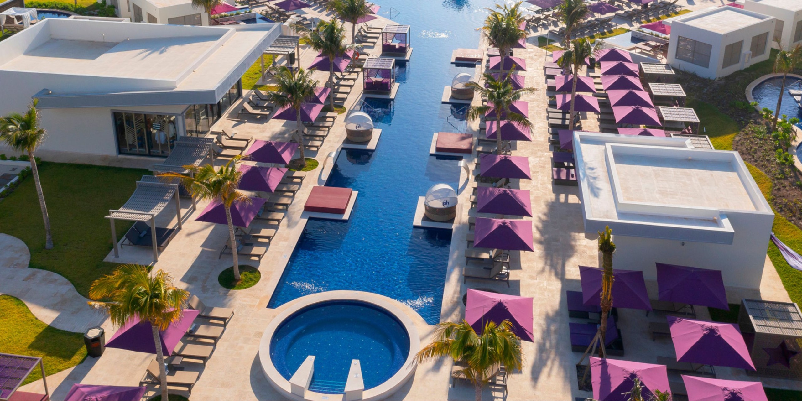 Aerial view of Planet Hollywood Adult Scene All Suites Cancun, a luxury resort featuring a long pool, surrounded by white buildings and purple umbrellas.