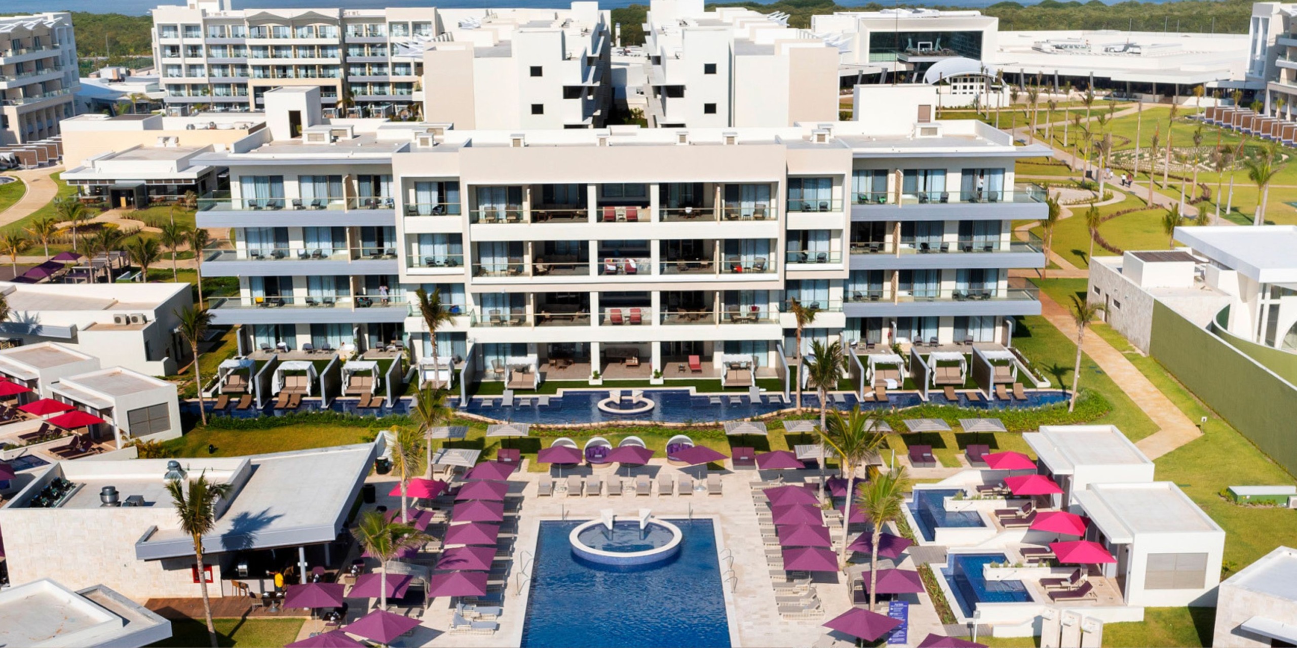 An aerial view of Planet Hollywood Adult Scene All Suites Cancun showcases its modern multi-story architecture, complete with a pool adorned by purple umbrellas in the foreground, all encircled by beautifully landscaped gardens.