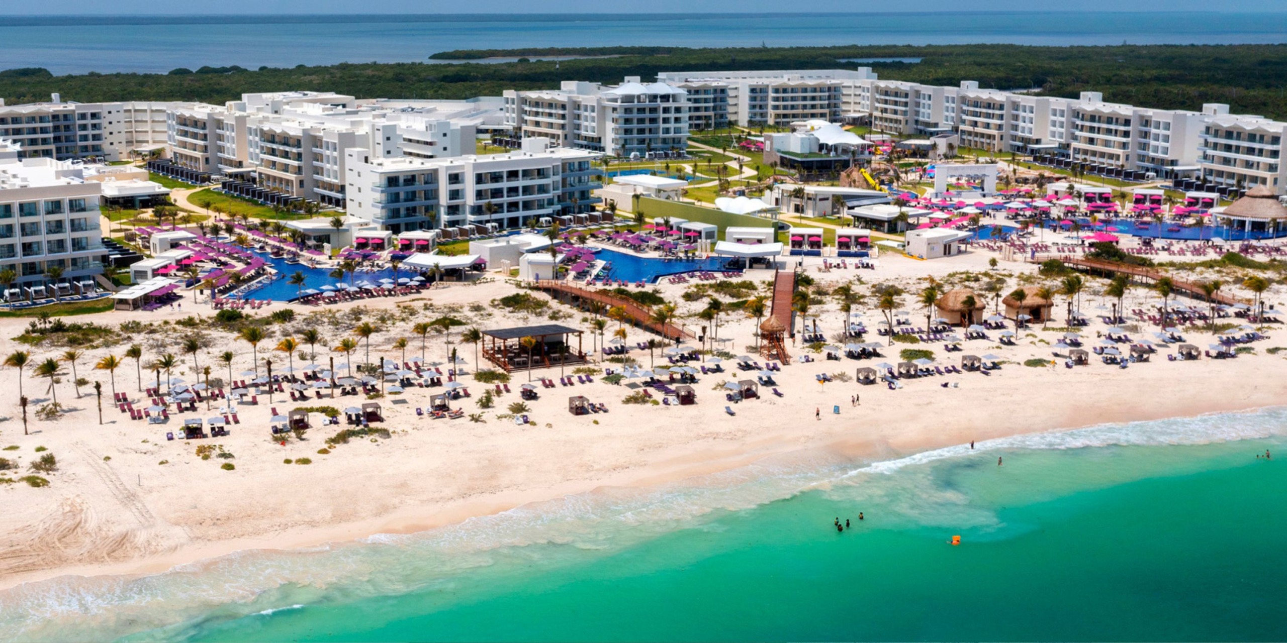 Aerial view of Planet Hollywood Adult Scene All Suites Cancun, a beachfront resort featuring white buildings, multiple pools, and vibrant pink-colored areas. The beach is lined with lounge chairs overlooking the turquoise ocean.