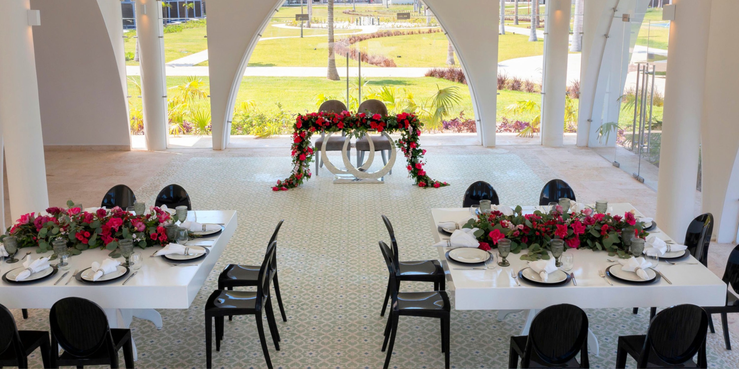 A wedding setup featuring two tables adorned with pink and red flowers, elegant white plates, and stylish black chairs, set against the backdrop of a floral archway with sweeping outdoor views of Planet Hollywood Adult Scene All Suites Cancun.