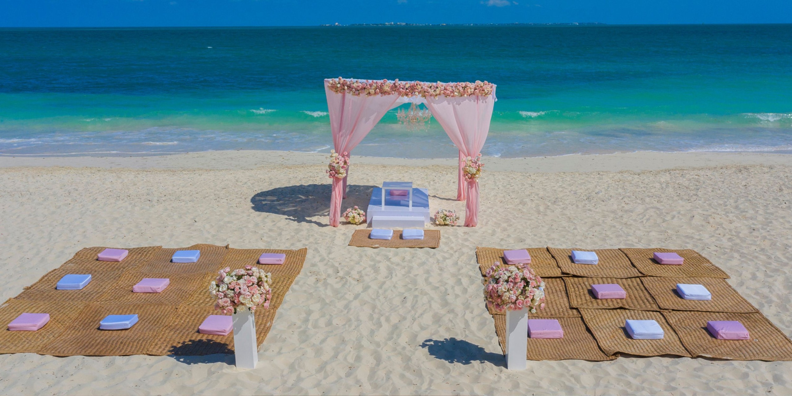Experience a stunning beach wedding setup at Planet Hollywood Adult Scene All Suites Cancun, featuring a pink floral arch, comfortable seating mats, and plush pillows on the sand, all perfectly arranged to face the breathtaking ocean.