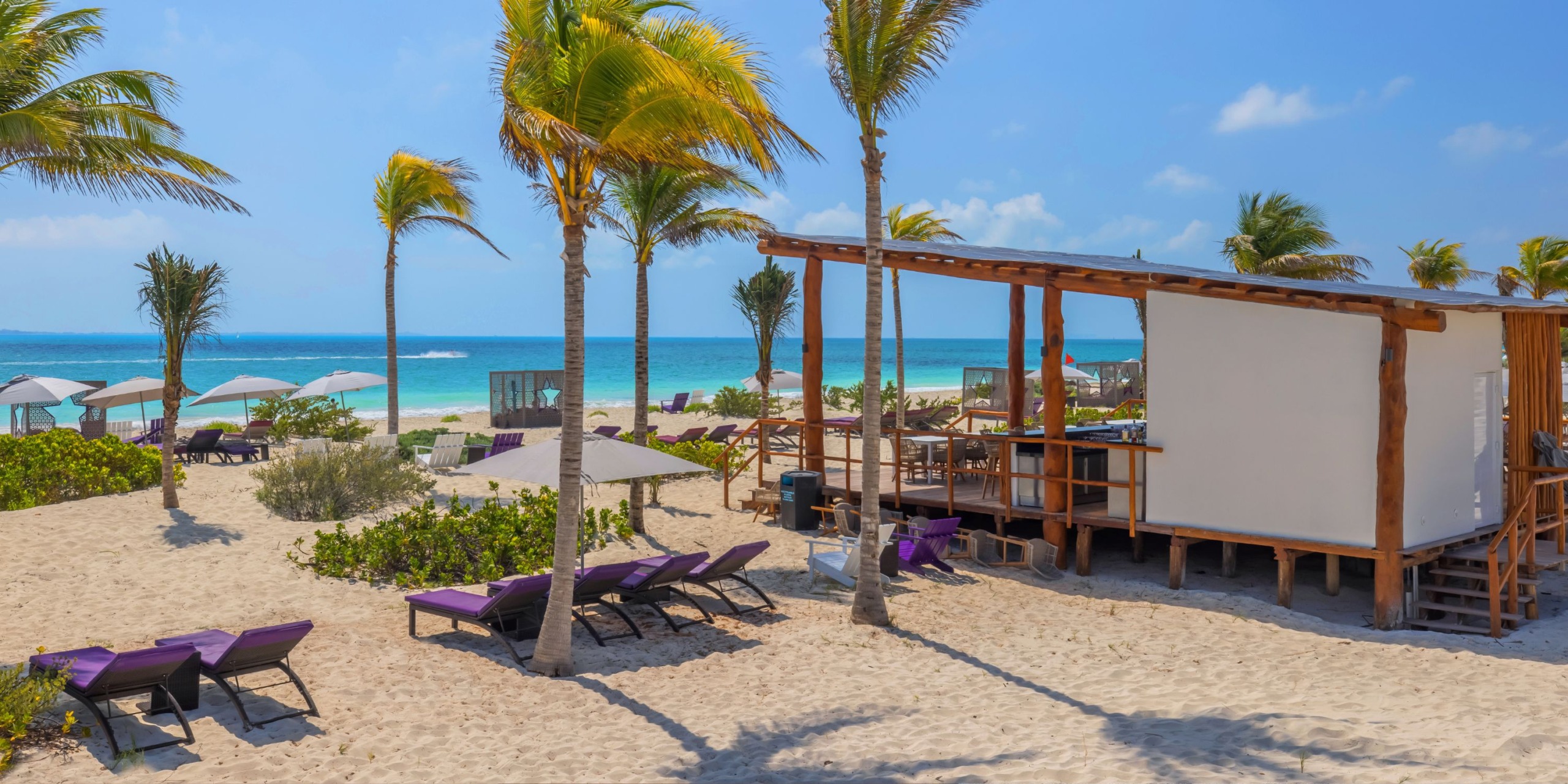 Experience the captivating scene at Planet Hollywood Adult Scene All Suites Cancun, where you'll find lounge chairs and palm trees against a backdrop of a wooden structure, all set on white sand with the blue sea under a clear sky.