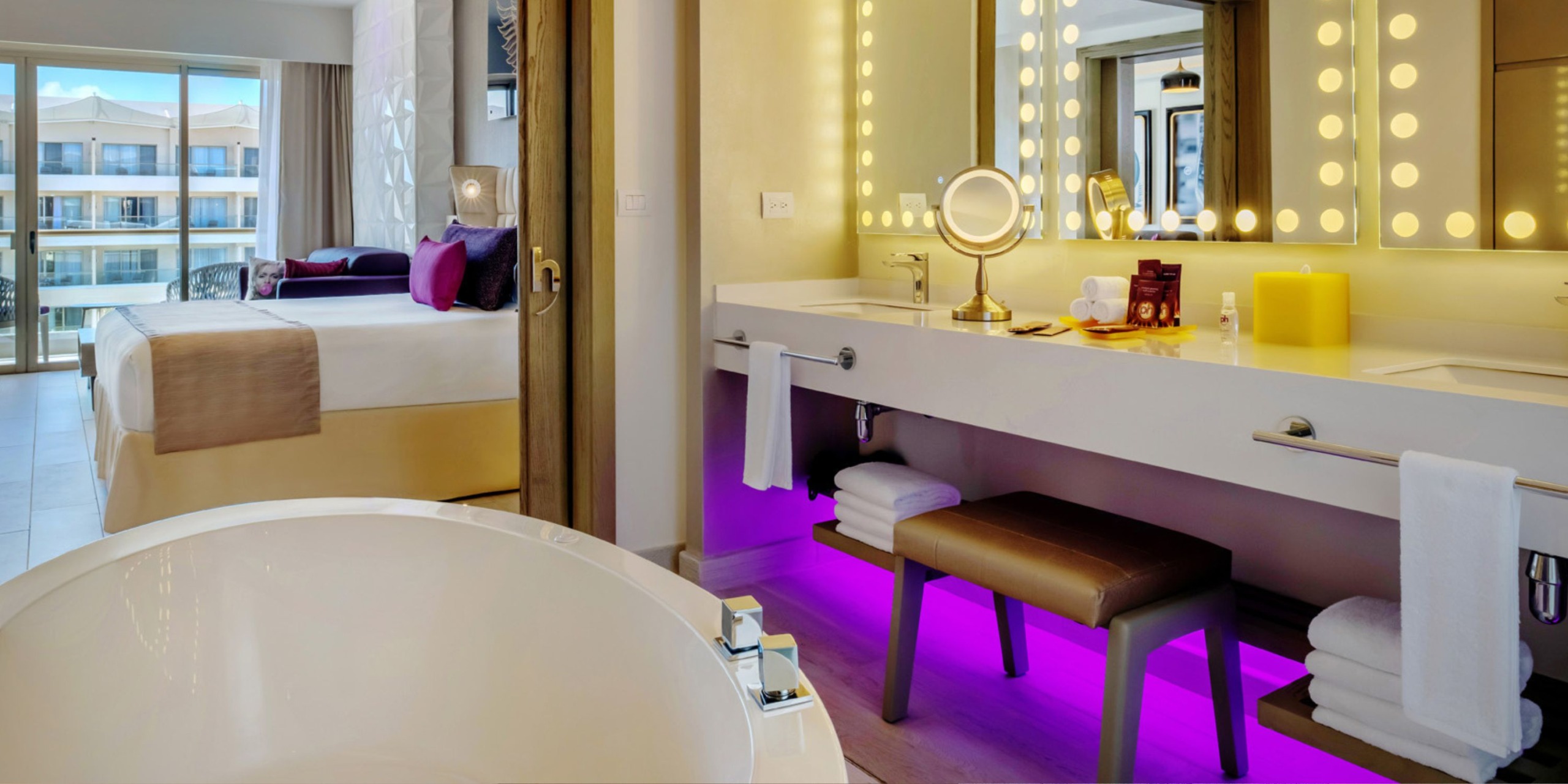 Experience a luxurious stay at Planet Hollywood Adult Scene All Suites Cancun, featuring a modern hotel bathroom with a spacious bathtub, an illuminated vanity mirror, and soothing purple ambient lighting beneath the counter. In the background, the bedroom with its plush bed opens up to an inviting balcony.