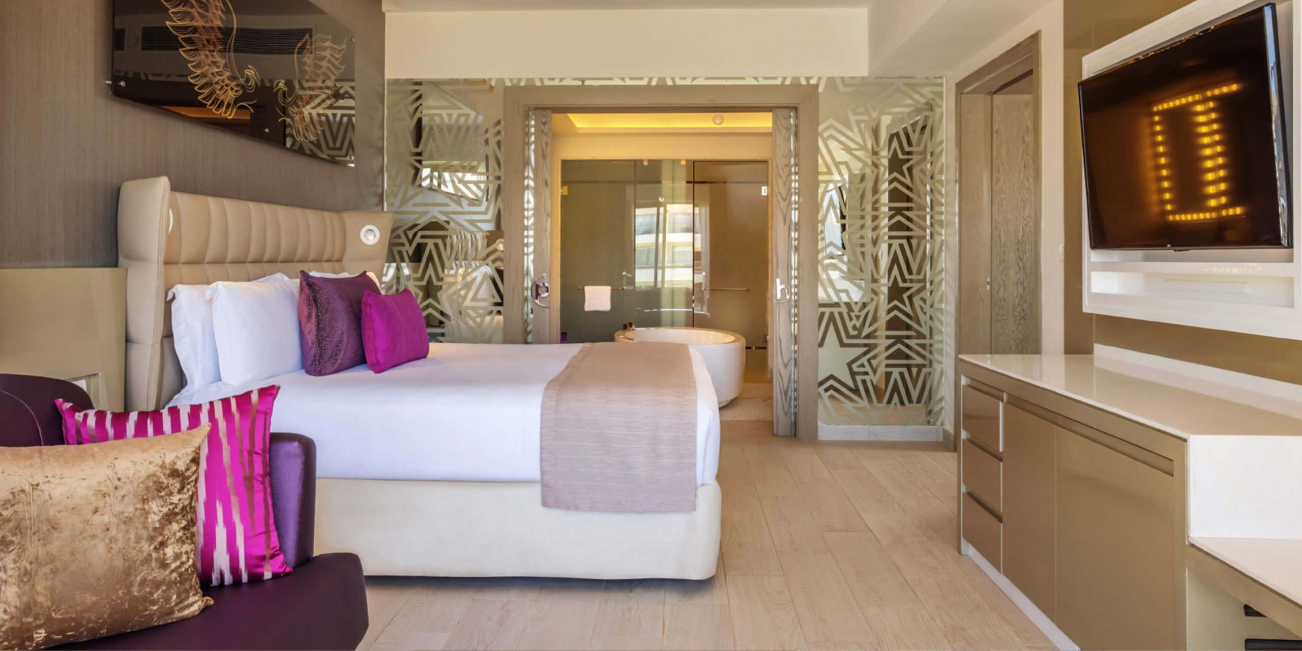 Enjoy a modern suite at the Planet Hollywood Adult Scene All Suites Cancun, featuring a spacious bed adorned with purple and gold accent pillows, a wall-mounted TV, and a bathroom view through an elegant glass partition.