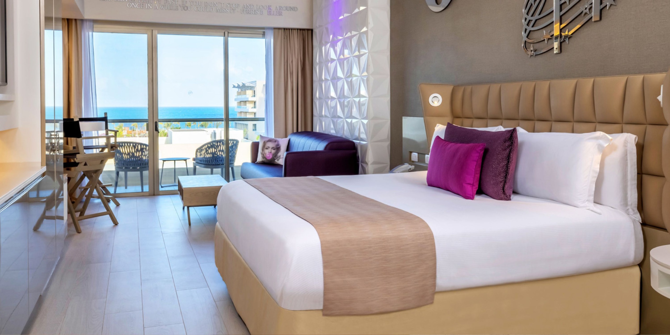 Experience luxury with a suite at Planet Hollywood Adult Scene All Suites Cancun, featuring a spacious bed, stylish purple sofa, and expansive floor-to-ceiling windows leading to a balcony showcasing breathtaking ocean views. The room boasts modern decor with pristine white walls and elegant light wood flooring.
