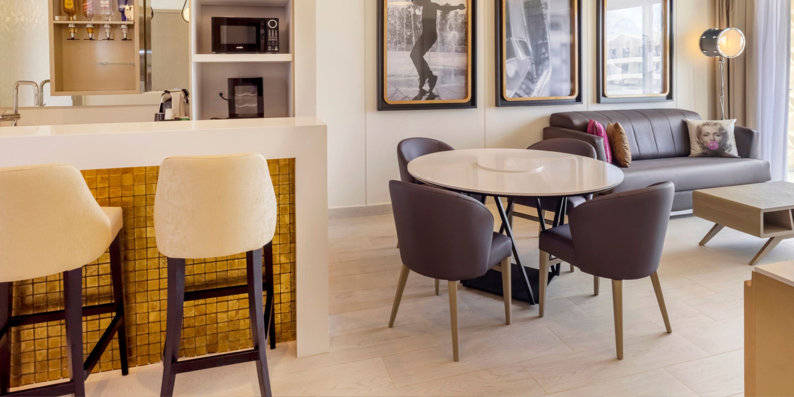 Discover contemporary elegance at the Planet Hollywood Adult Scene All Suites Cancun, featuring a living area complete with a round dining table and six chairs, complemented by a chic gray sofa and sophisticated framed wall art. In the foreground, you'll find a countertop accompanied by stylish bar stools, while a sleek kitchenette provides the perfect backdrop.