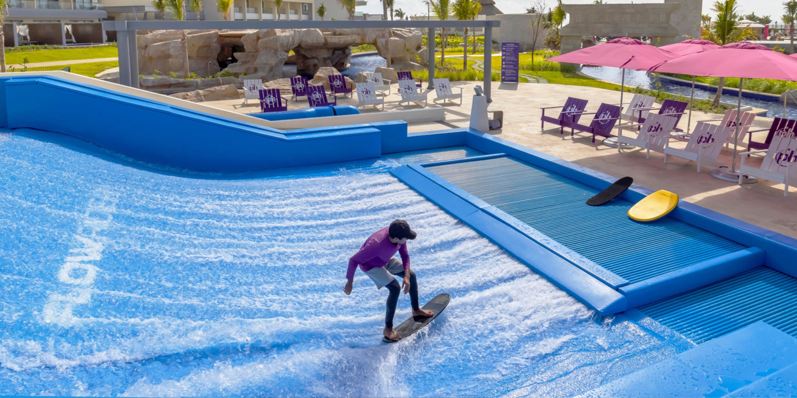 A person wearing a purple shirt is surfing on a blue artificial wave machine at the Planet Hollywood Adult Scene All Suites Cancun, while two surfboards are placed on a platform surrounded by lounge chairs and umbrellas.