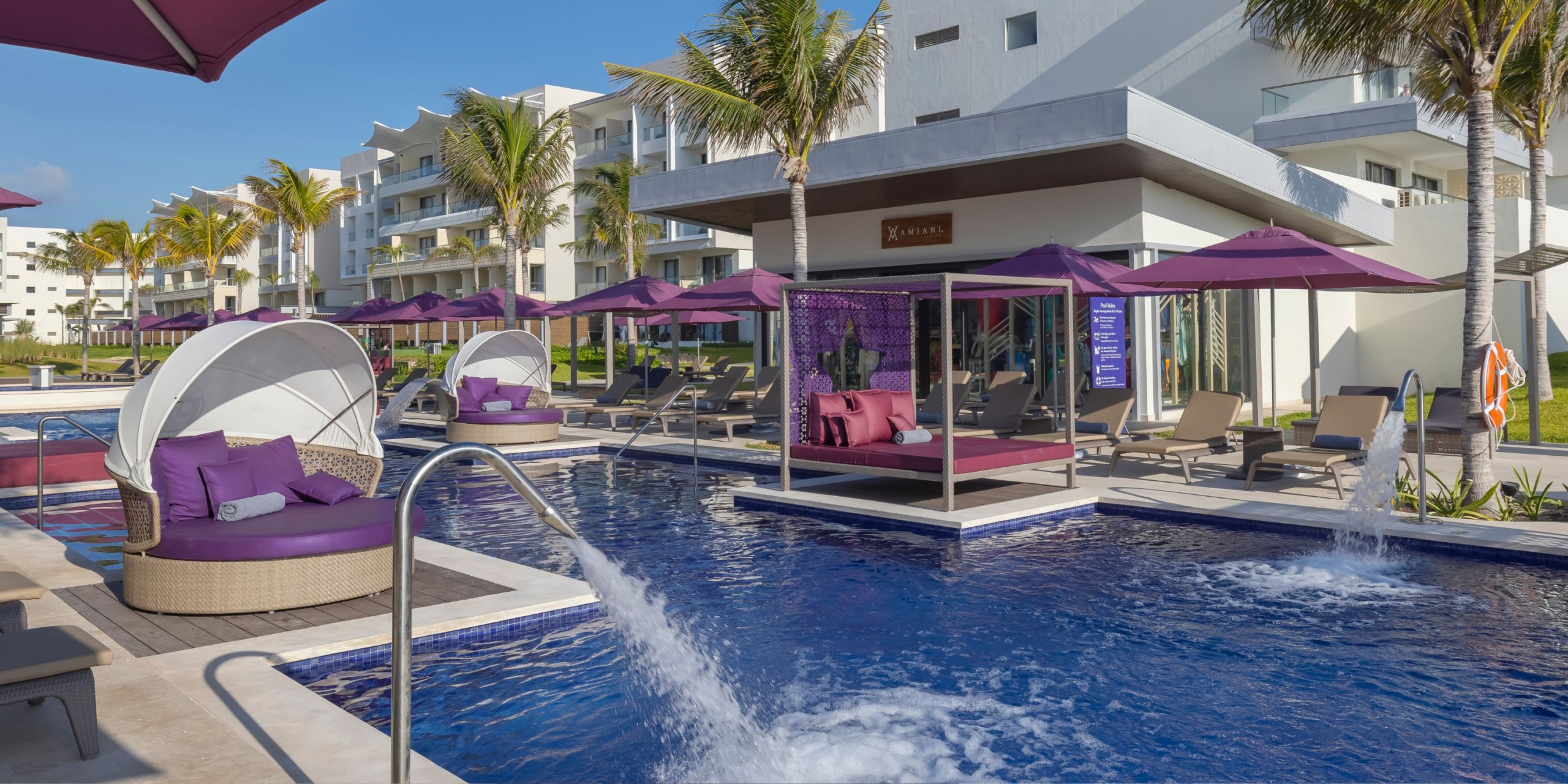 The Planet Hollywood Adult Scene All Suites Cancun offers a chic pool area featuring lounge chairs and cabanas, surrounded by palm trees and pristine white structures. The seating is highlighted with purple umbrellas and cushions, while water jets gracefully spray into the pool.