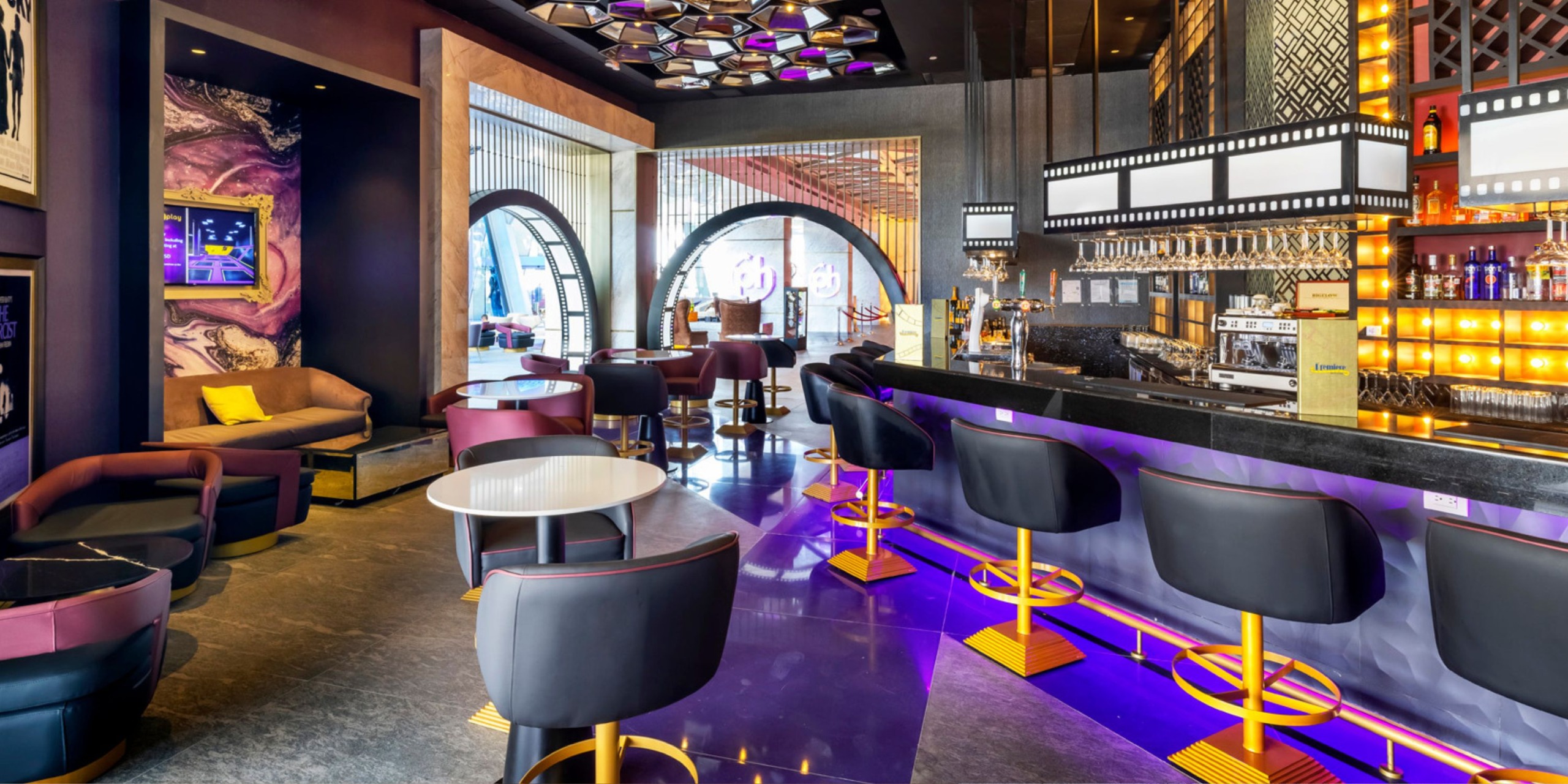 The interior of Planet Hollywood Adult Scene All Suites Cancun features a modern bar with purple and gold accents, curved bar stools, tables, and arched windows. It is brightly lit with patterned ceilings and showcases a diverse selection of bottles on display.