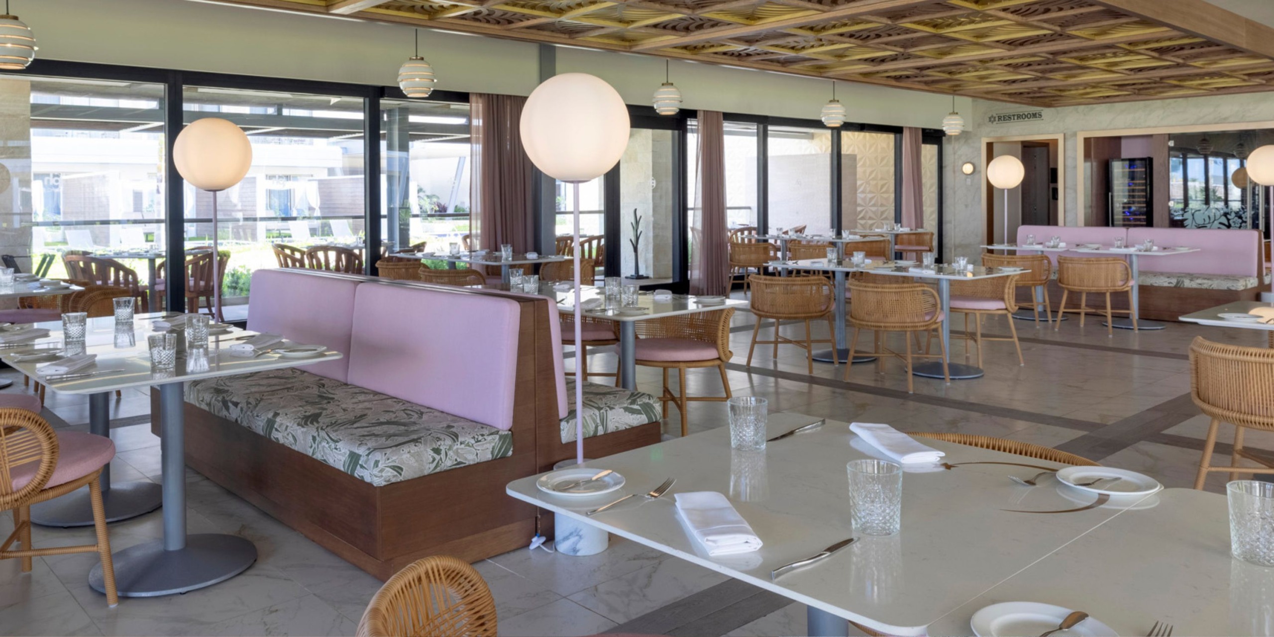 A stylish space in Planet Hollywood Adult Scene All Suites Cancun features empty tables with pink cushioned seating and woven chairs, illuminated by globe pendant lights. Large windows along one wall provide an abundance of natural light.