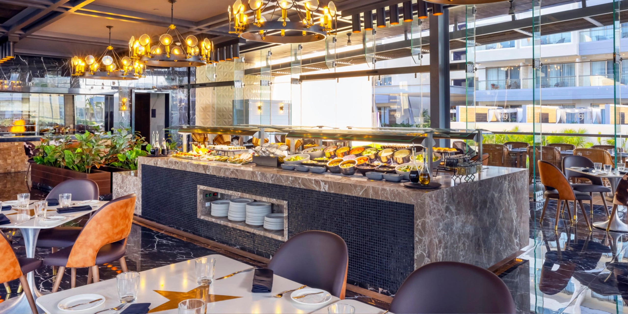 The Planet Hollywood Adult Scene All Suites Cancun features a contemporary buffet area adorned with marble counters, a variety of dishes, and refined lighting. The tables and chairs are strategically placed around the room to offer a delightful view of the outdoors.