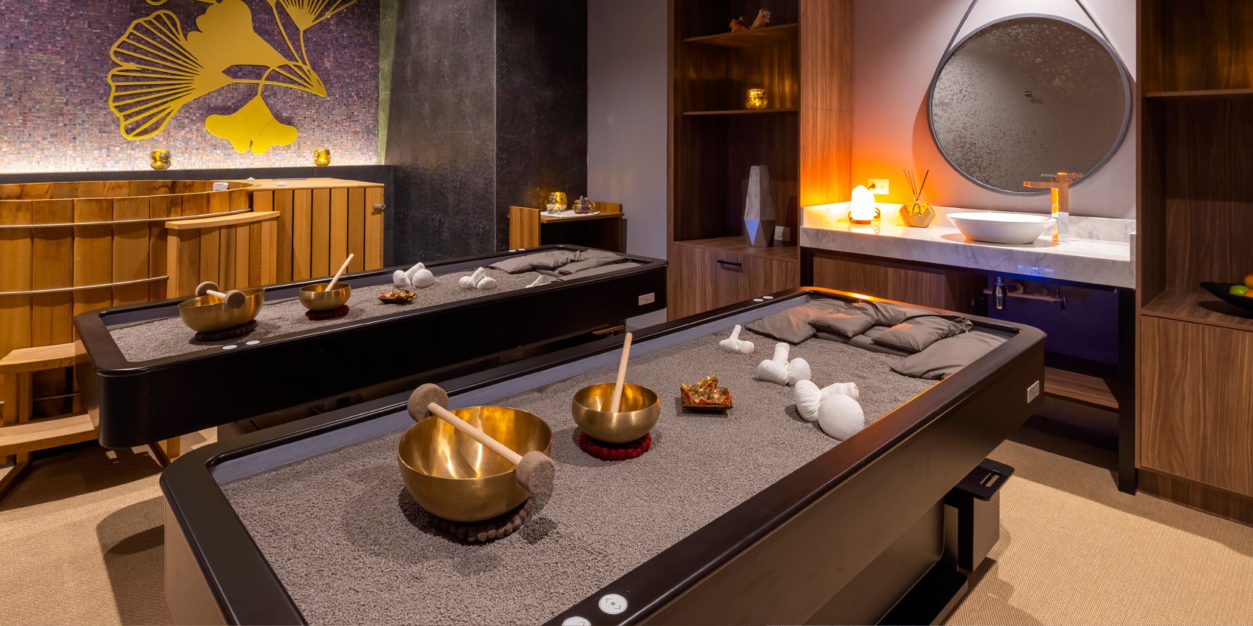 A tranquil space at the Planet Hollywood Adult Scene All Suites Cancun includes two sand massage tables, a sink adorned with a round mirror, and decorative touches such as golden bowls and neatly folded towels.