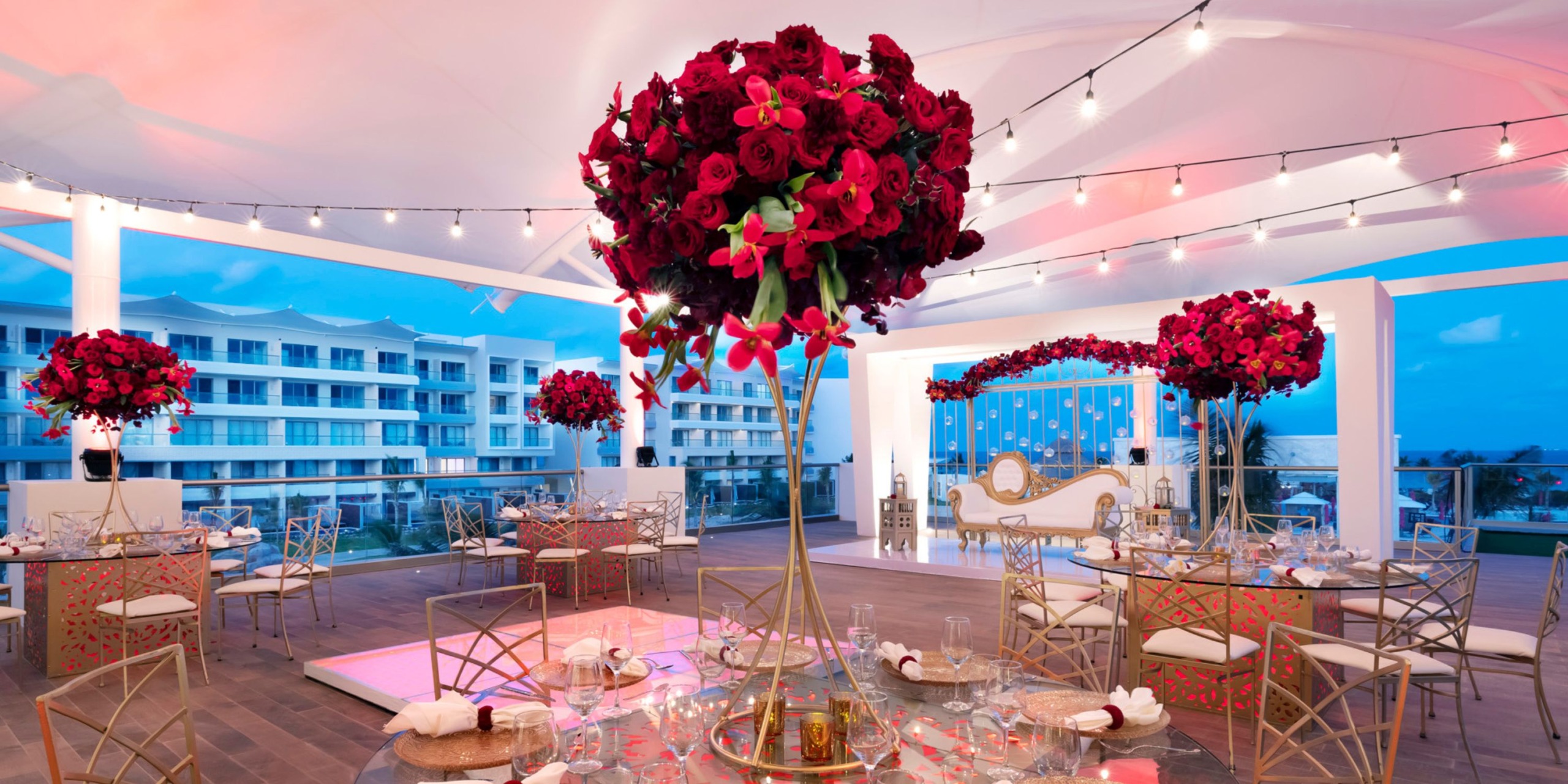 Experience the sophisticated atmosphere at Planet Hollywood Adult Scene All Suites Cancun, featuring tall floral centerpieces with rich red hues, elegant gold chairs, and enchanting string lights. The venue boasts striking white architecture and offers a stunning view of modern cityscapes in the background.