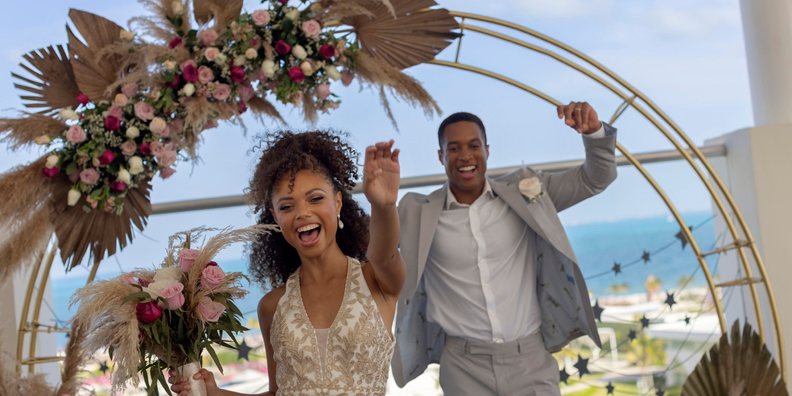 A joyful couple celebrates under a floral arch by the sea at Planet Hollywood Adult Scene All Suites Cancun. The woman holds a bouquet, and both are smiling with their arms raised.