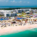 Aerial view of the Planet Hollywood Adult Scene All Suites Cancun resort, featuring white buildings, palm trees, and loungers on the sand. The turquoise ocean and lush greenery provide a stunning background.