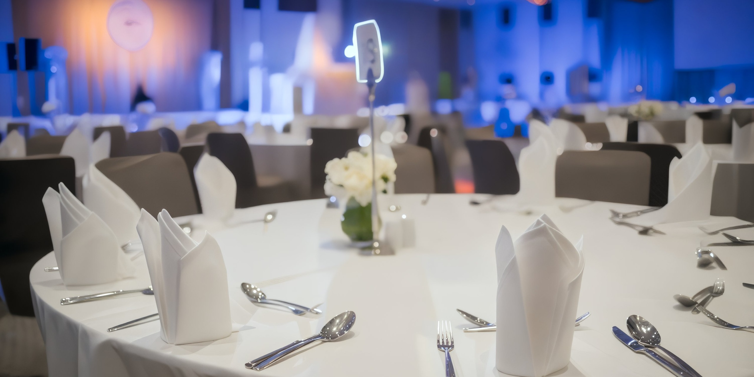 The Princess Grand Jamaica banquet hall is elegantly arranged for an event, with round tables adorned in white tablecloths, neatly folded napkins, and polished cutlery. Each table is beautifully accented with a small flower centerpiece.