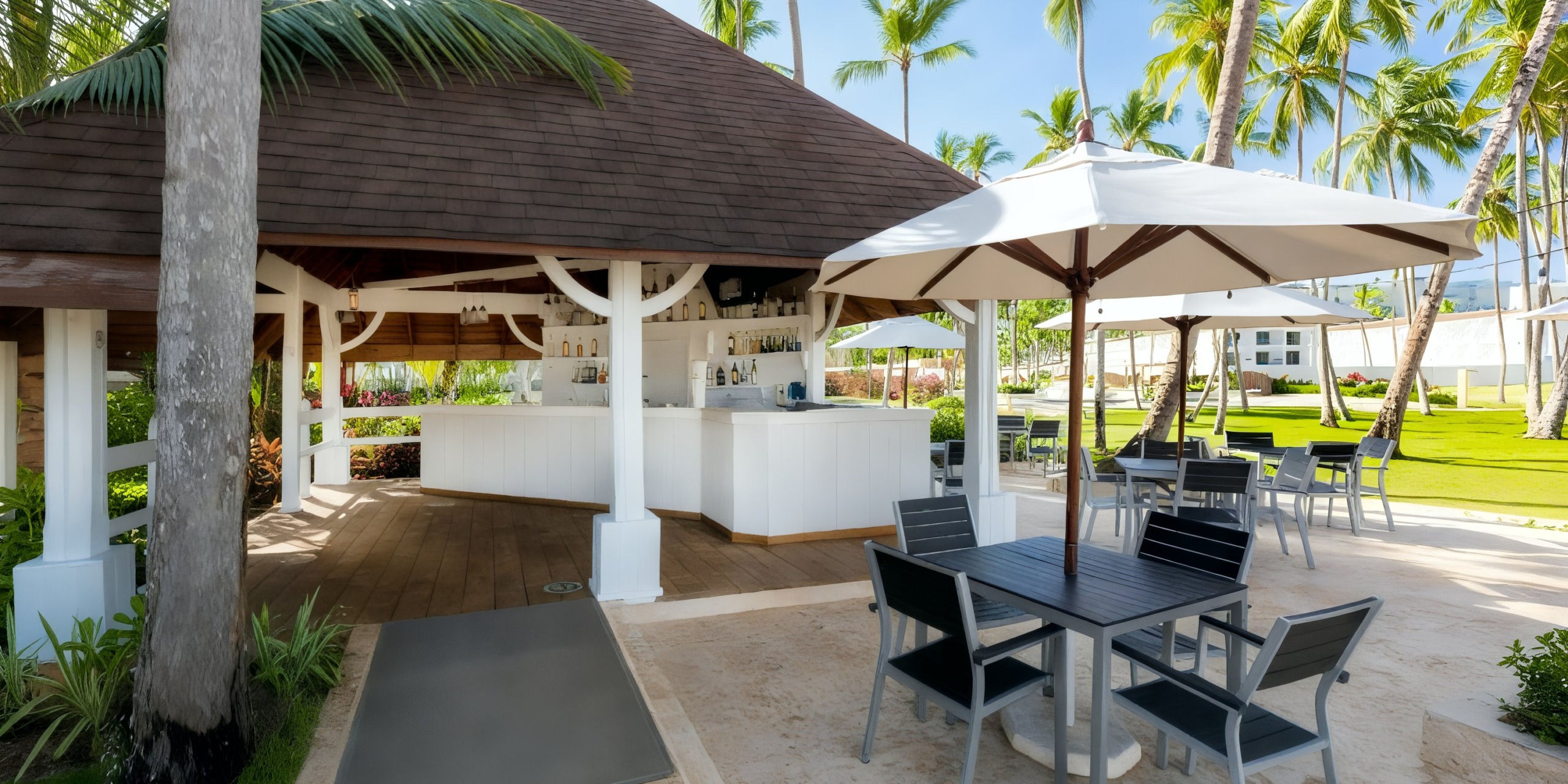 Viva V Samana by Wyndham features an outdoor bar with a wooden roof, nestled among palm trees and accompanied by patio tables equipped with umbrellas on a sunny day.