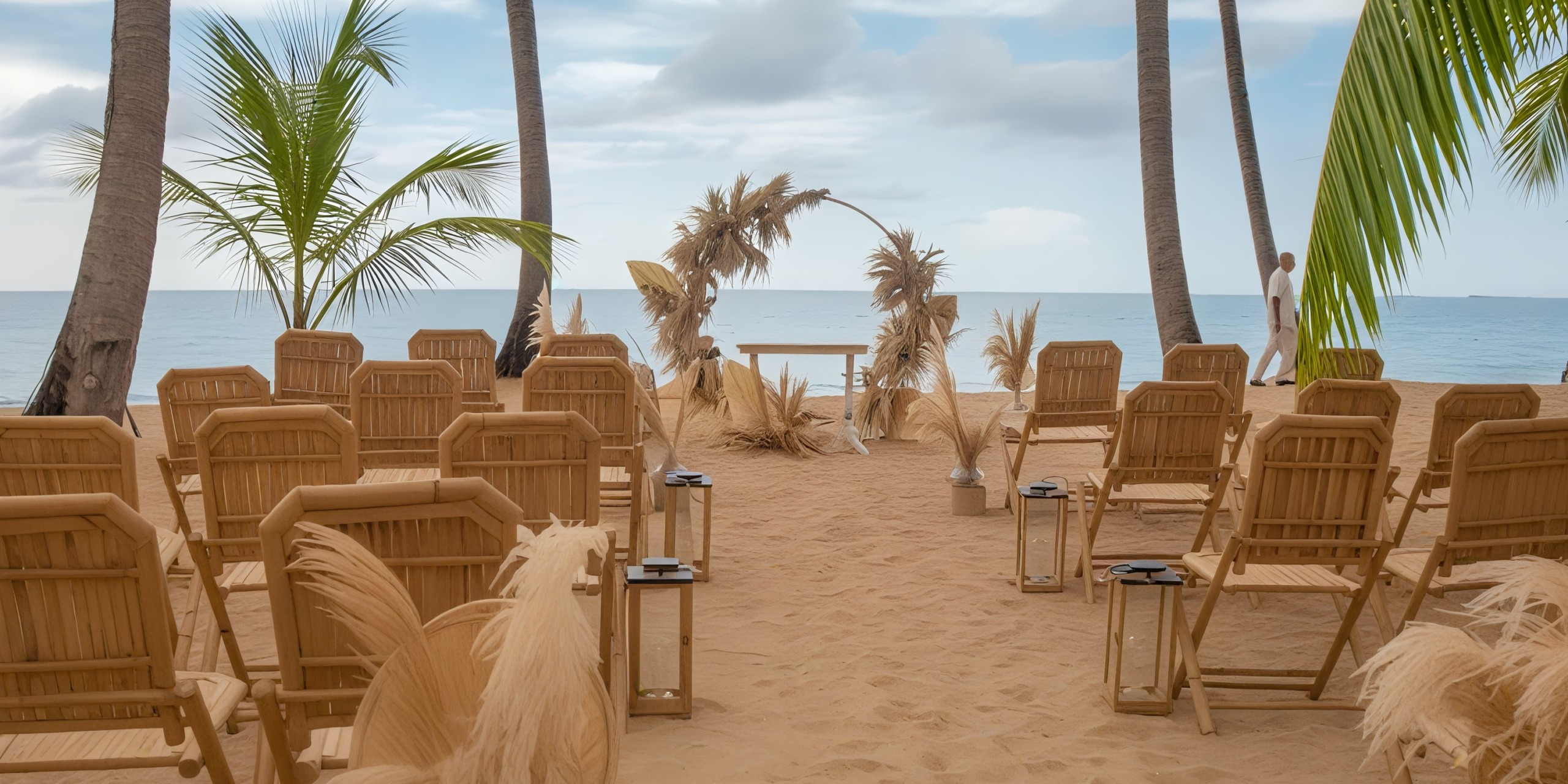 Experience a breathtaking beach wedding setup at Viva V Samana by Wyndham, featuring wooden chairs, a dried floral arch, and lanterns adorning the sand. The picturesque scene is complete with an ocean backdrop under a partly cloudy sky.