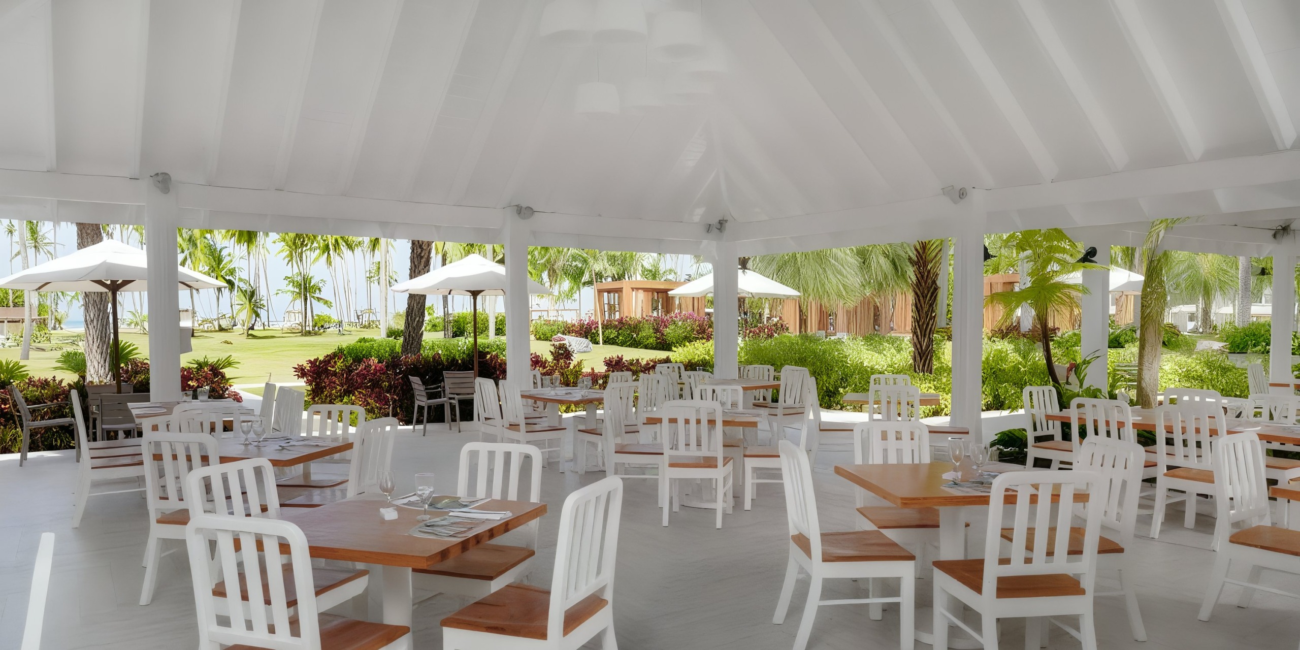 The Viva V Samana by Wyndham features an outdoor dining area adorned with white wooden tables and chairs, beautifully surrounded by palm trees and lush greenery.