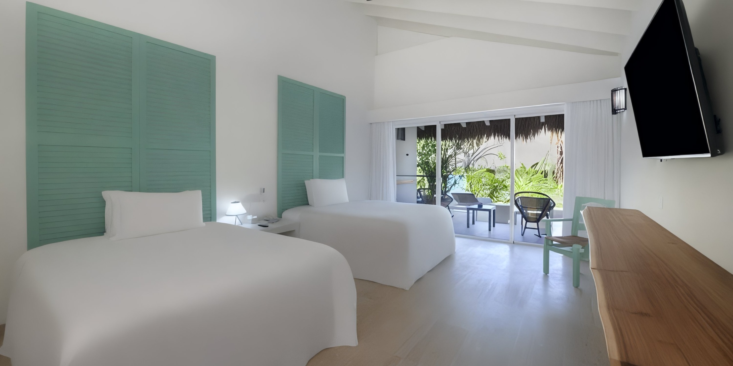 The Viva V Samana by Wyndham hotel room includes two double beds, a wall-mounted TV, and a wooden desk. It also offers a sliding glass door that opens to a small terrace with chairs, providing views of lush tropical plants.